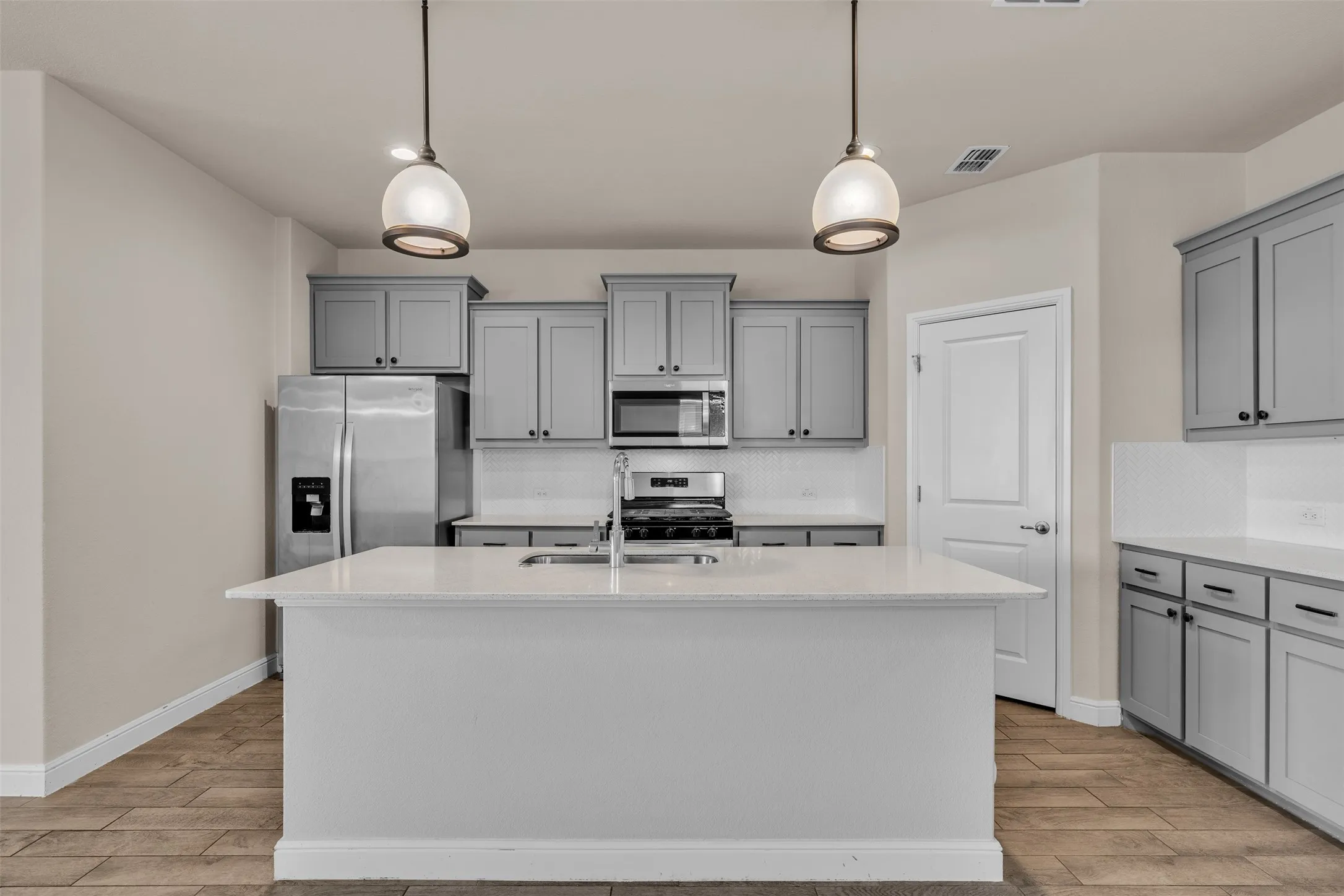 Kitchen featuring a central island with an integrated sink, pendant lighting, stainless steel appliances, grey cabinetry, and white countertops