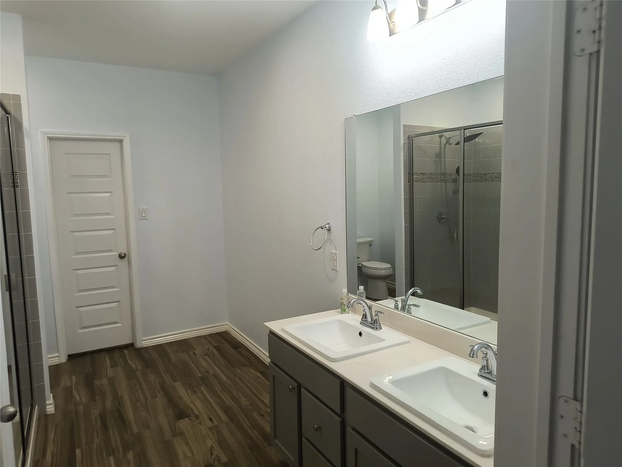 Double vanity with integrated sinks, wood-finish flooring, a frameless mirror, and a glass-enclosed shower with tiled walls