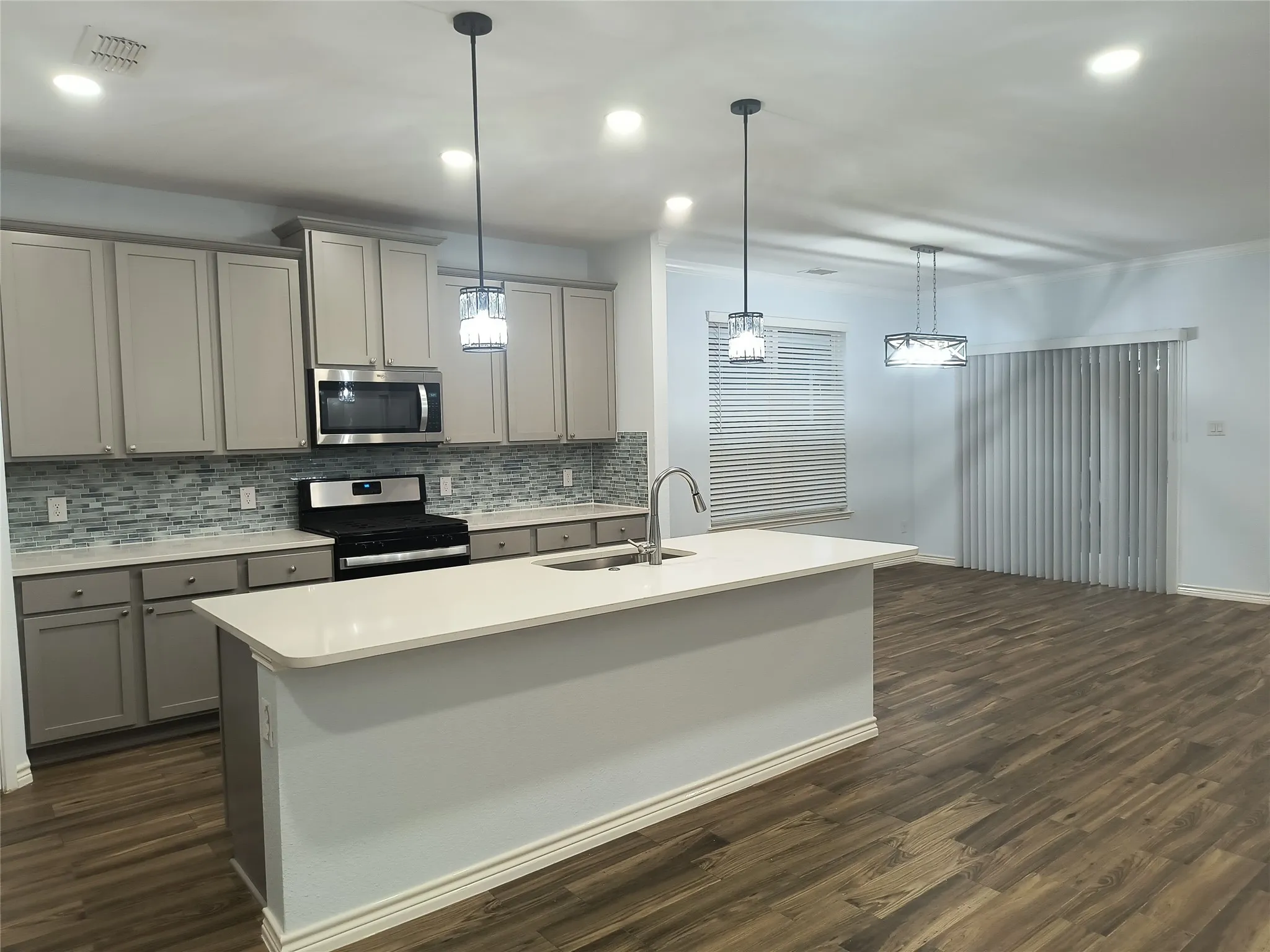 Open-concept kitchen featuring gray shaker cabinetry, a white countertop island with an integrated sink, a mosaic tile backsplash, stainless steel appliances, and recessed lighting