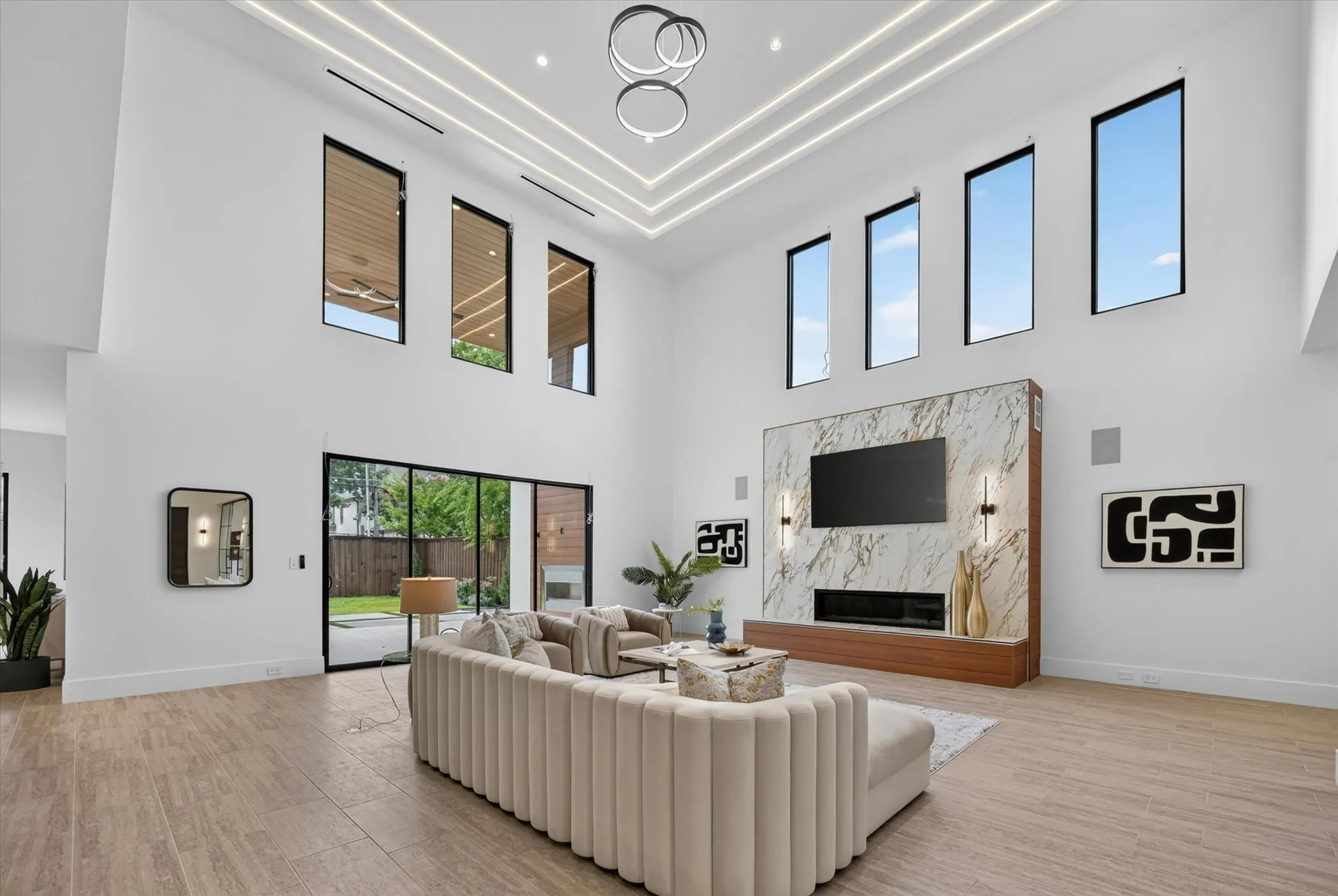 Double-height ceiling with integrated linear lighting, large format tile flooring, floor-to-ceiling sliding glass doors, and a stone-clad fireplace with built-in wood-finish shelving