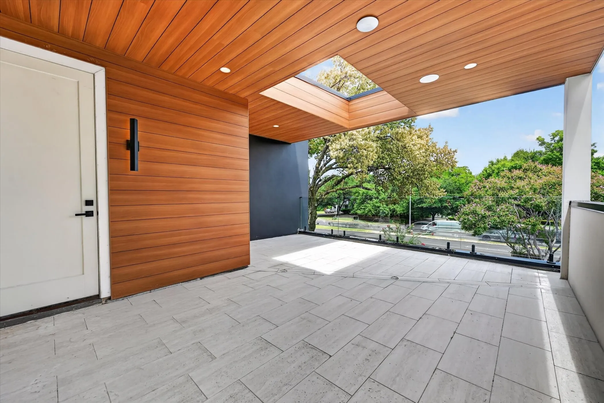 Spacious outdoor balcony featuring patterned tile flooring, wood-paneled ceiling, integrated skylight, and a sleek glass railing