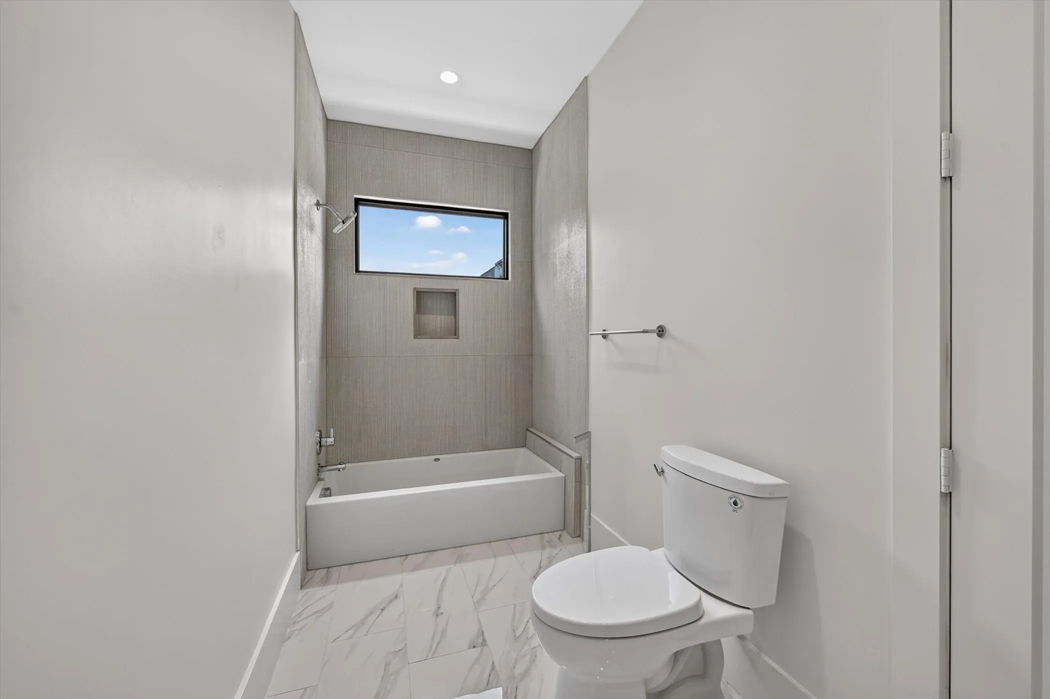 Bathroom featuring a rectangular soaking tub with a built-in shower niche, a horizontal window, tile-finish wall surround, and marble-finish tile flooring
