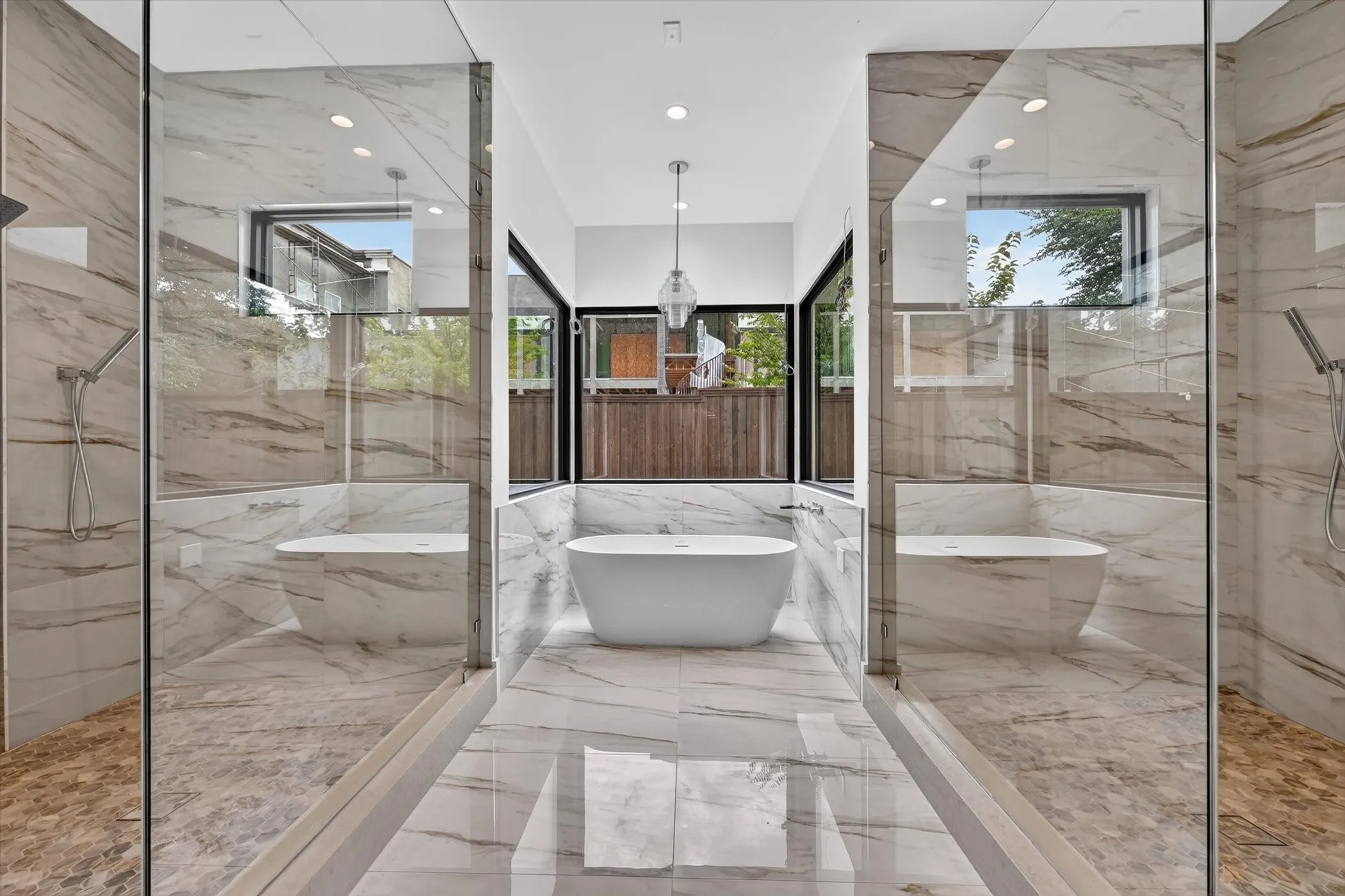 Freestanding soaking tub, dual glass-enclosed showers with marble-look tile, and recessed lighting fixtures