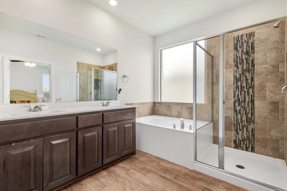 Bathroom featuring a dual vanity with dark wood cabinetry, a large frameless mirror, a built-in soaking tub, a separate glass-enclosed shower with tile surround and decorative accent, and wood-finish flooring