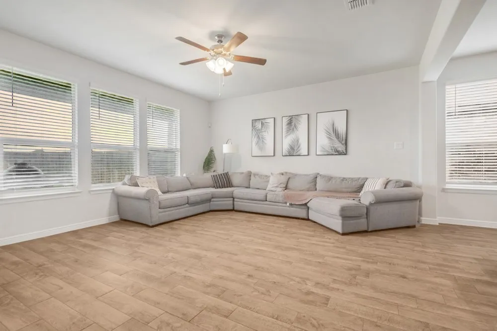 Spacious room featuring wood-finish flooring, multiple large windows with blinds, a ceiling fan with integrated lighting, and neutral wall tones