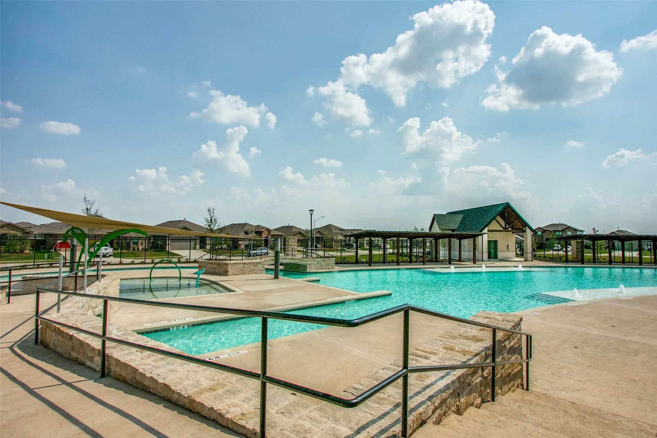 Expansive community swimming pool with integrated fountains, a separate shallow splash pad area with water features, a shaded pergola structure, and a clubhouse with a gabled roof