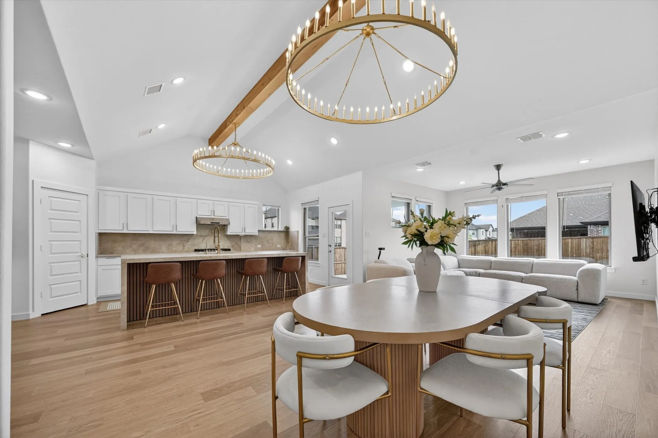 Open-concept living space featuring a vaulted ceiling with exposed wood beam, two circular modern chandeliers, and light wood-finish flooring