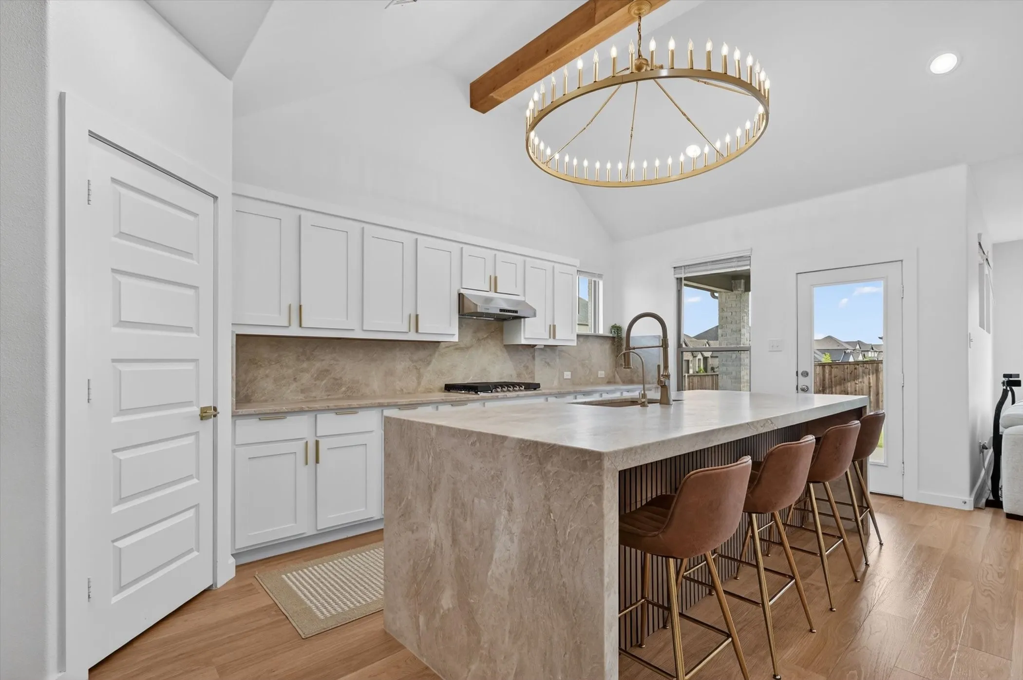 Modern kitchen featuring a large waterfall island, white cabinetry with gold-finish hardware, wood-finish flooring, a stone backsplash, and a vaulted ceiling with a decorative wood beam and chandelier