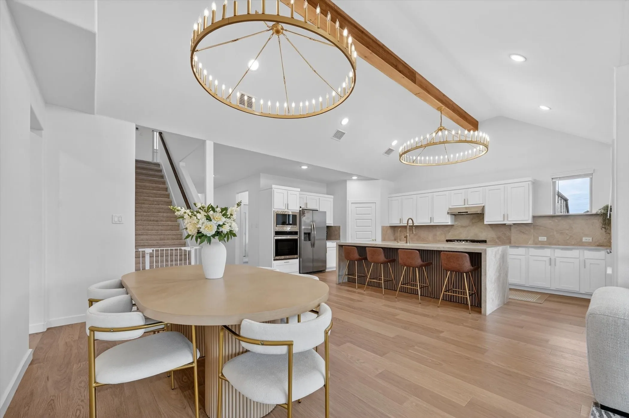 Open-concept living space featuring wood-finish flooring, a vaulted ceiling with exposed wood beam, and two large circular chandeliers