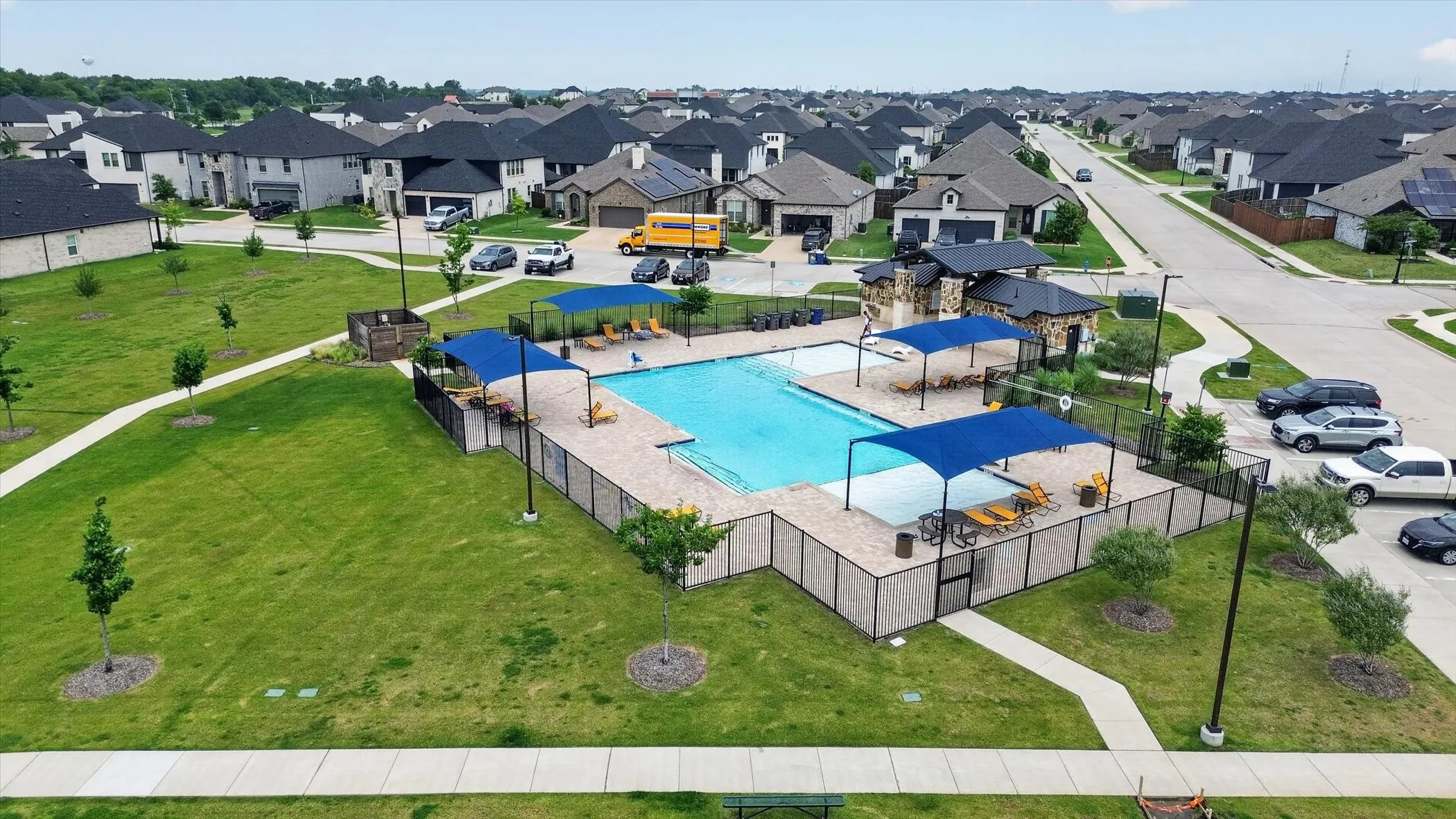 Community swimming pool featuring a perimeter fence, a stone-clad pool house, and multiple shaded lounge areas