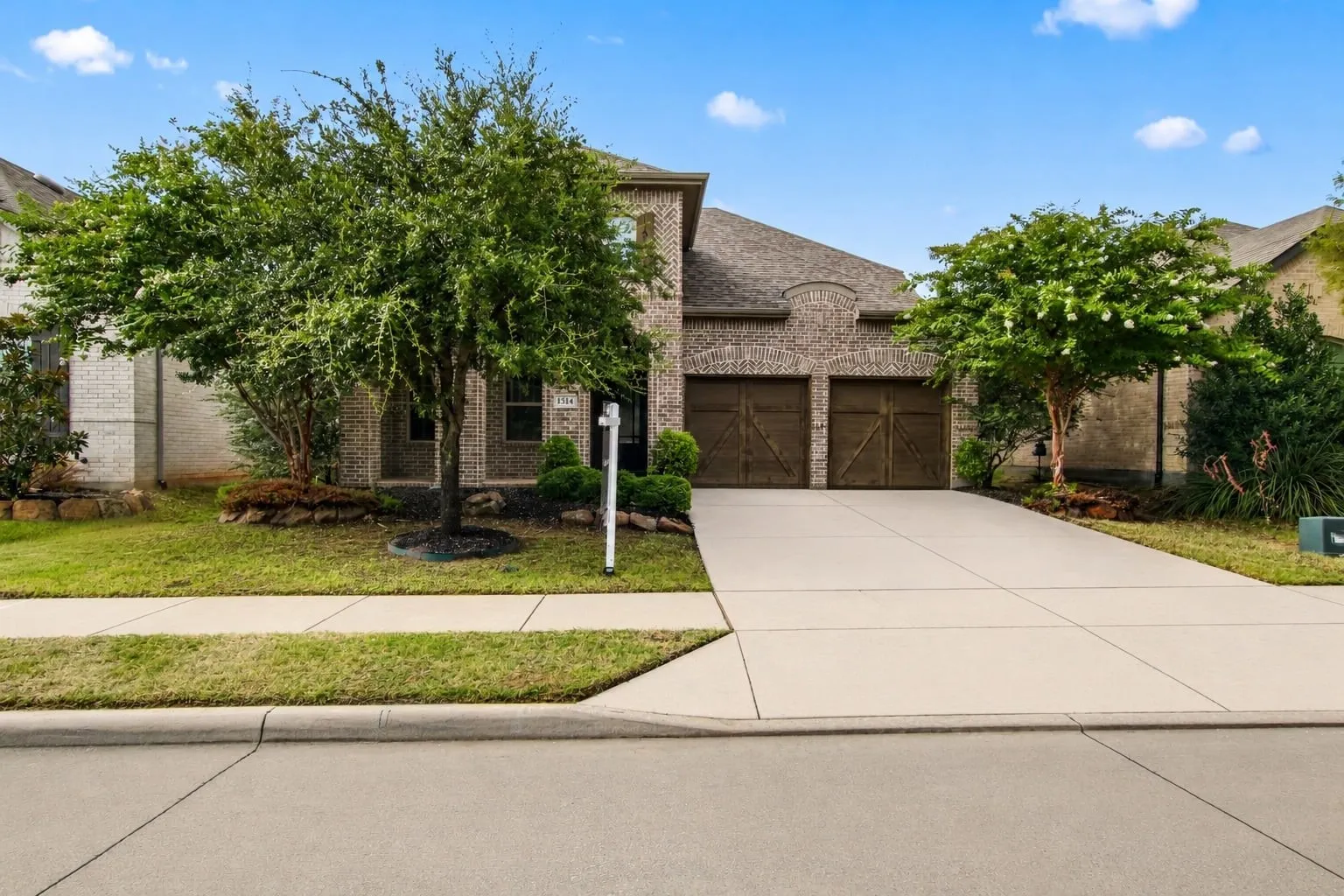 Brick facade home featuring a two-car garage with wood-finish doors, a concrete driveway, and a manicured lawn with mature trees and shrubbery