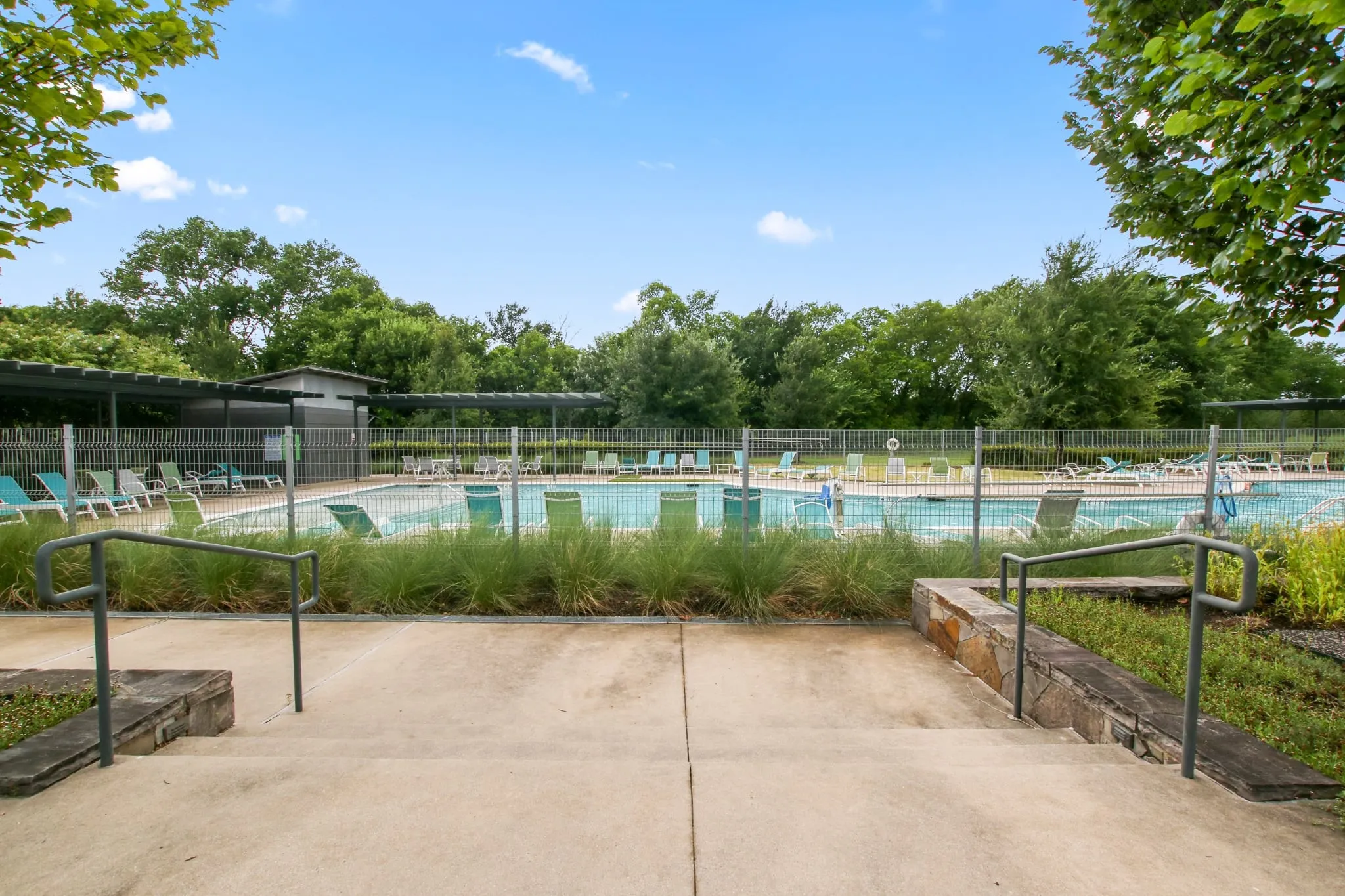 Community swimming pool featuring a perimeter fence, a shaded lounge area, and mature trees
