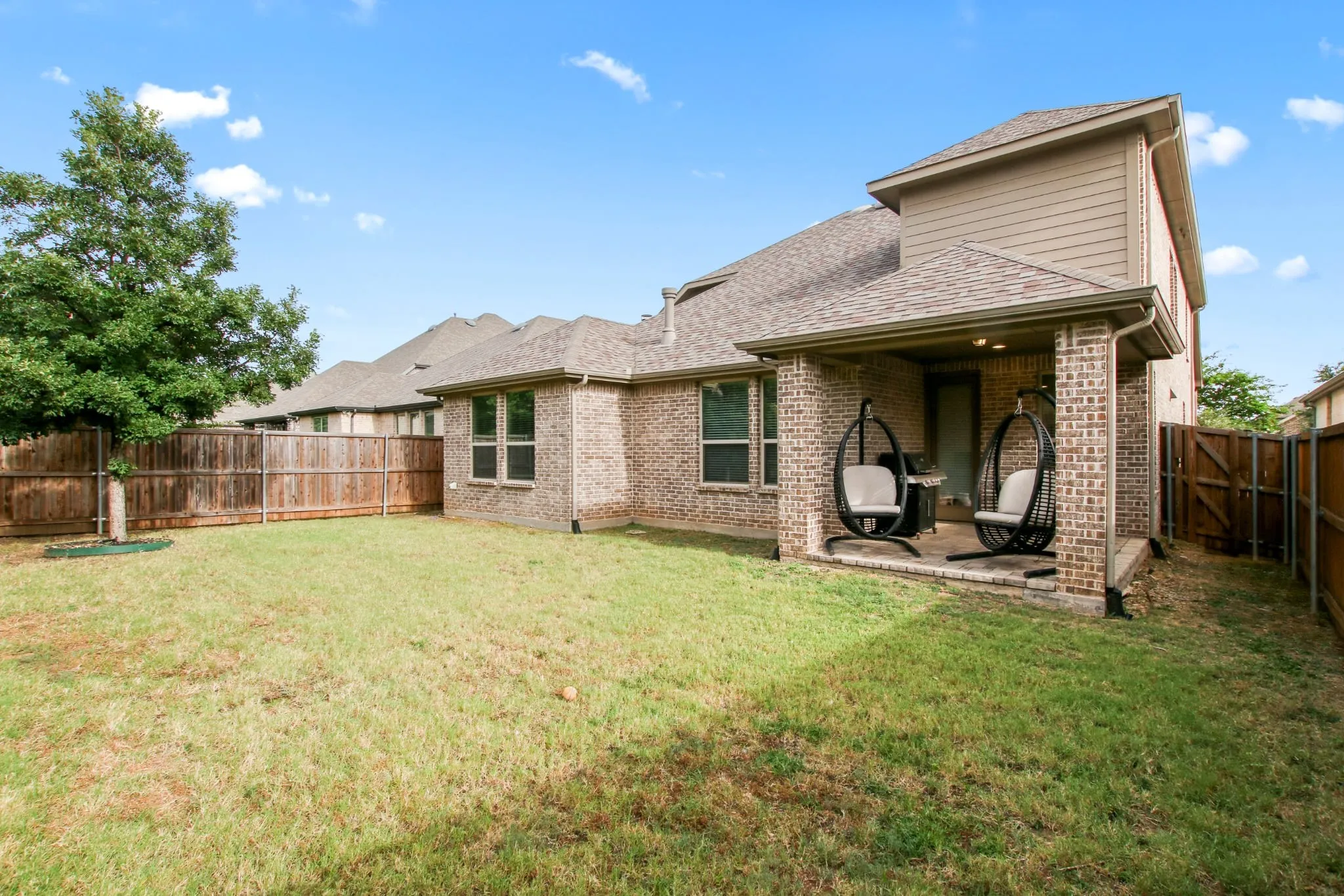 Expansive fenced backyard featuring a covered patio with brick columns, a manicured lawn, and mature trees