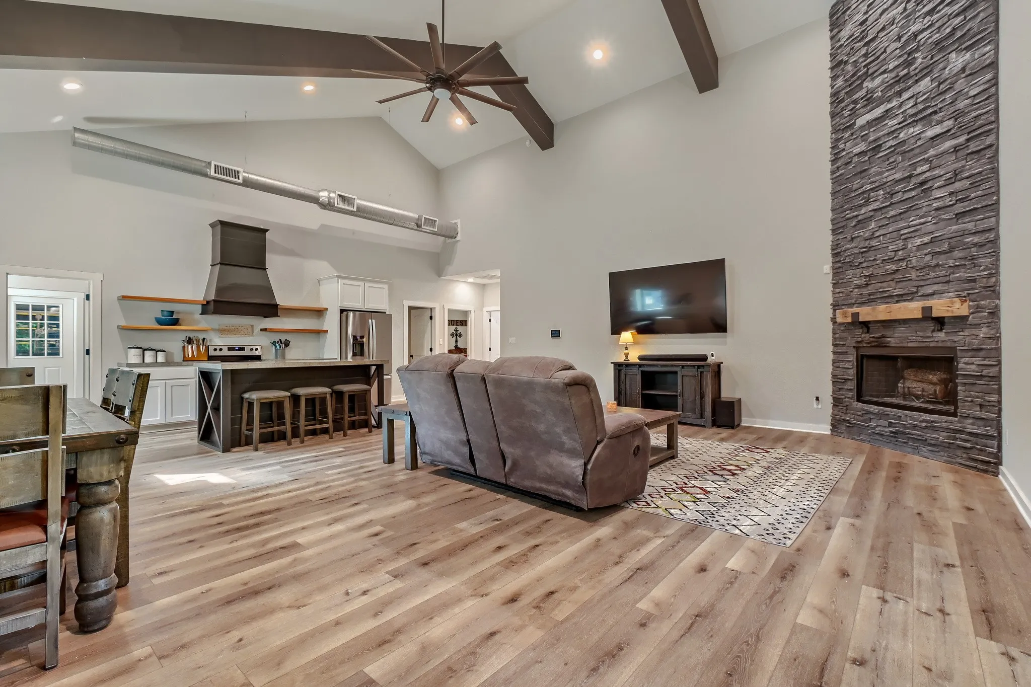 View of the open concept living, dining and kitchen upon entering, featuring vaulted ceilings with exposed beams, luxury vinyl wood flooring, floor to ceiling stone fireplace with a timber mantel.