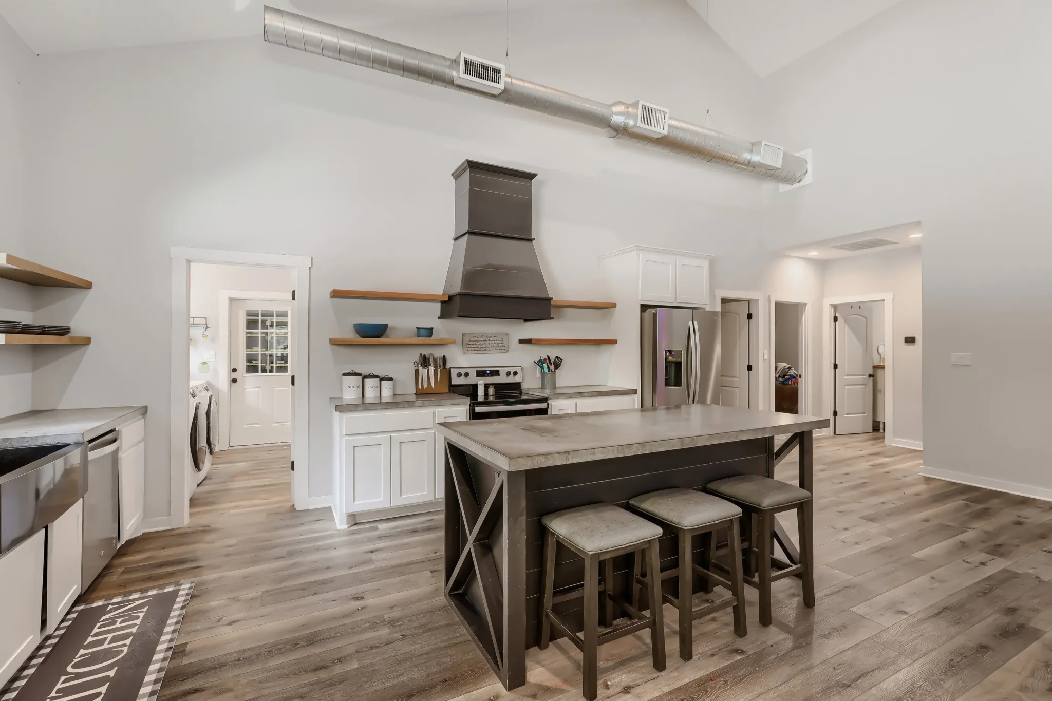 Open-concept living space featuring vaulted ceilings with exposed beams, luxury vinyl wood flooring, large kitchen island with concrete top, stainless steel appliances and large windows overlooking 5 private acres.