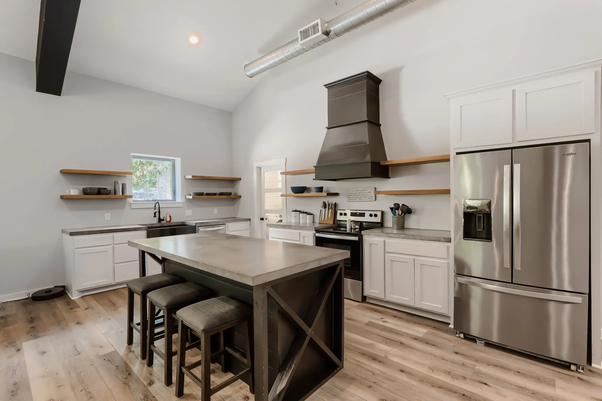 Open-concept living space featuring vaulted ceilings with exposed beams, luxury vinyl wood flooring, large kitchen island with concrete top, stainless steel appliances and large windows overlooking 5 private acres.
