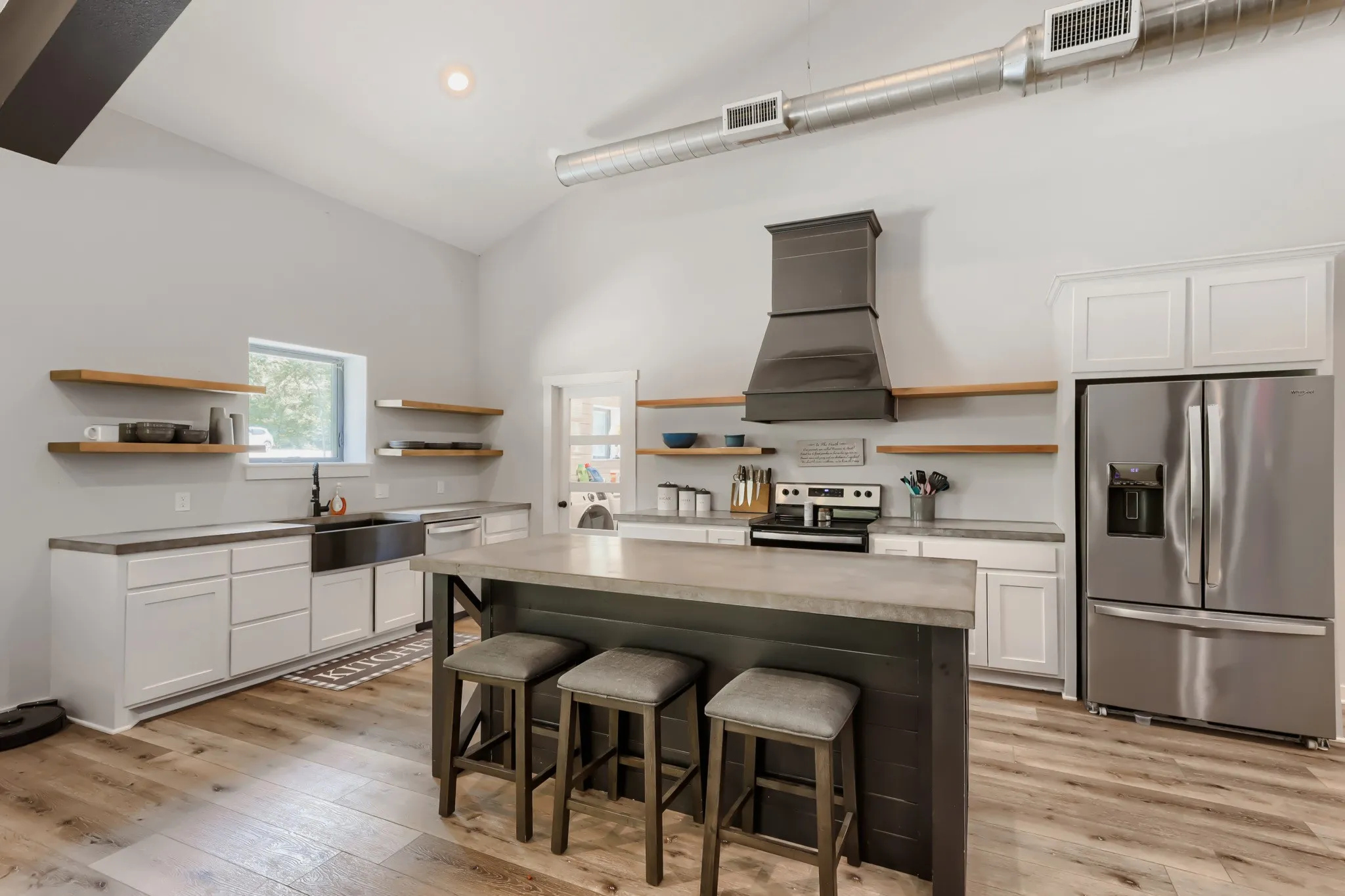 Open-concept living space featuring vaulted ceilings with exposed beams, luxury vinyl wood flooring, large kitchen island with concrete top, stainless steel appliances and large windows overlooking 5 private acres.