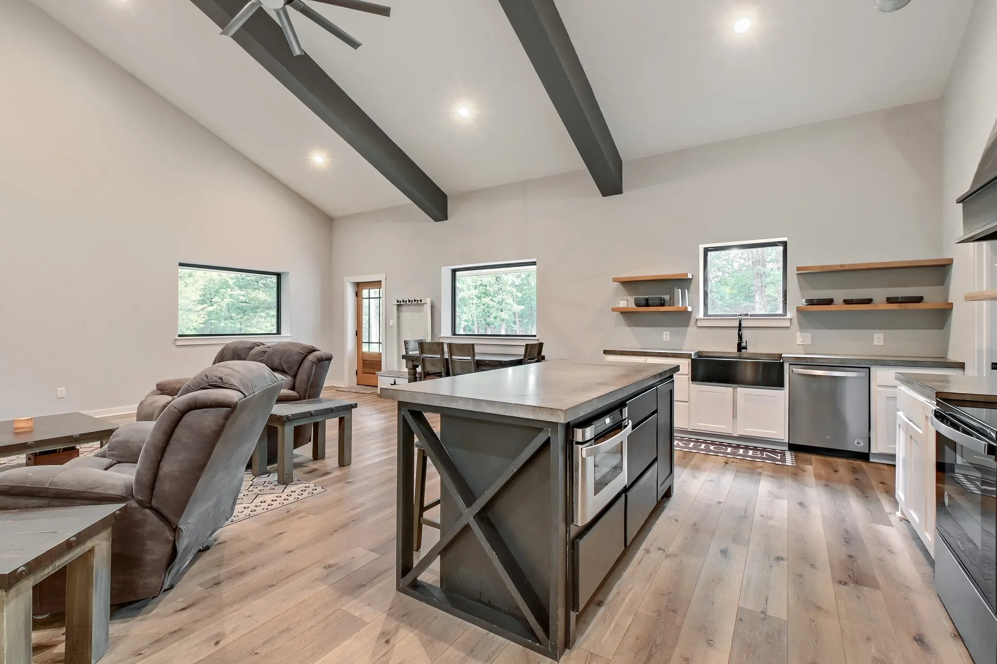 Open-concept living space featuring vaulted ceilings with exposed beams, luxury vinyl wood flooring, large kitchen island with concrete top, stainless steel appliances and large windows overlooking 5 private acres.