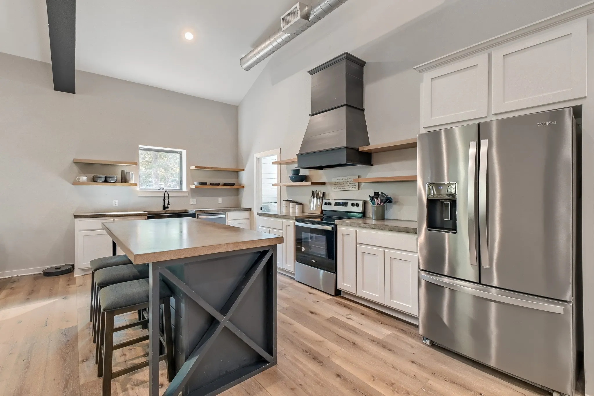 Spacious open farmhouse kitchen featuring a large, central eat-in island, stainless steel appliances, open wood shelving, white cabinetry, custom vent hood and luxury vinyl wood flooring.
