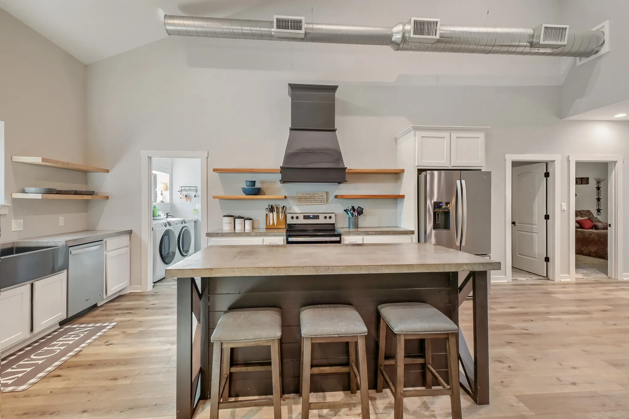 Spacious open farmhouse kitchen featuring a large, central eat-in island, stainless steel appliances, open wood shelving, white cabinetry, custom vent hood and luxury vinyl wood flooring.