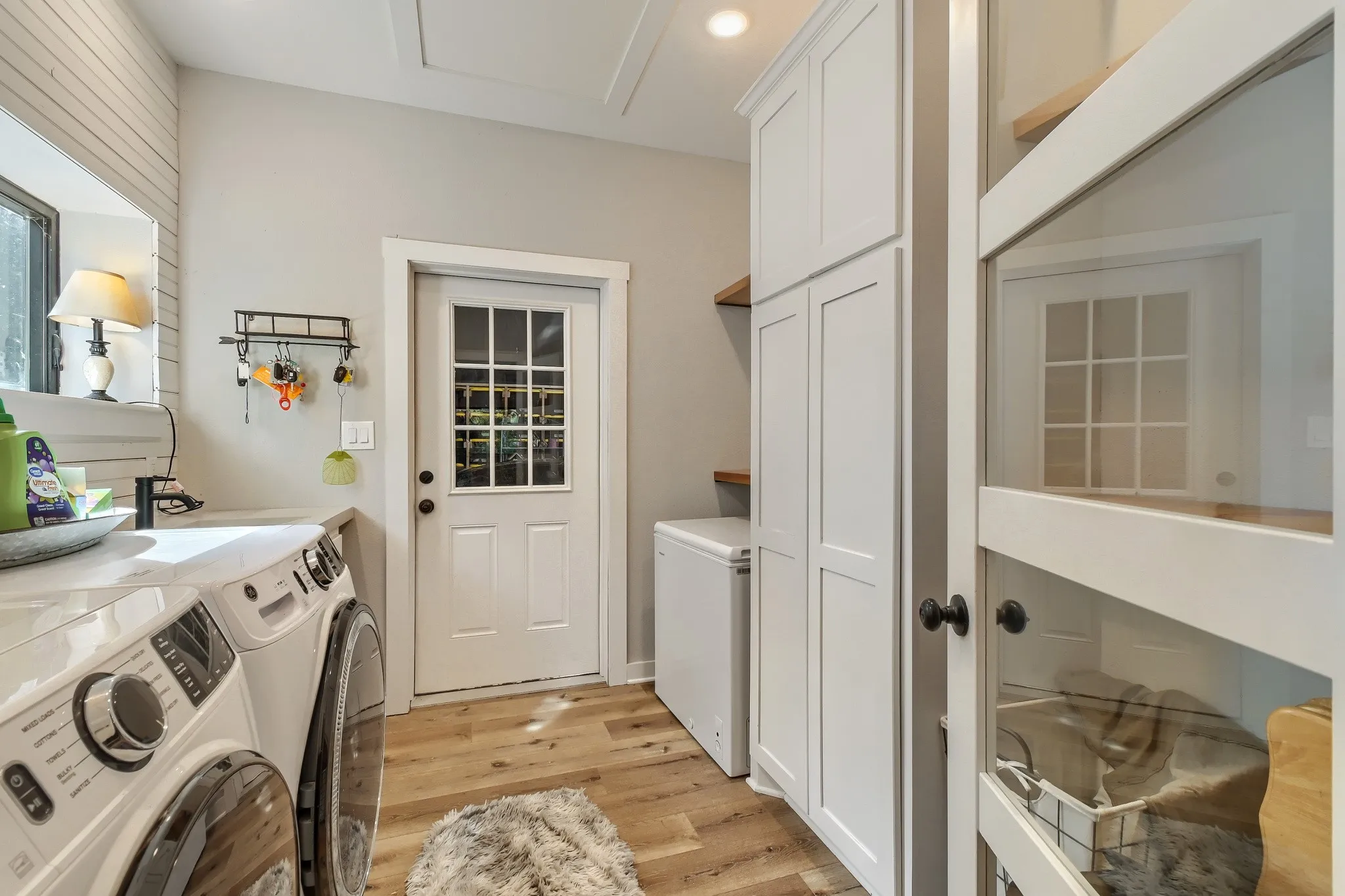 Dedicated farmhouse laundry, pantry and storage area featuring white cabinetry and a white paneled door with direct access to oversized garage/shop.