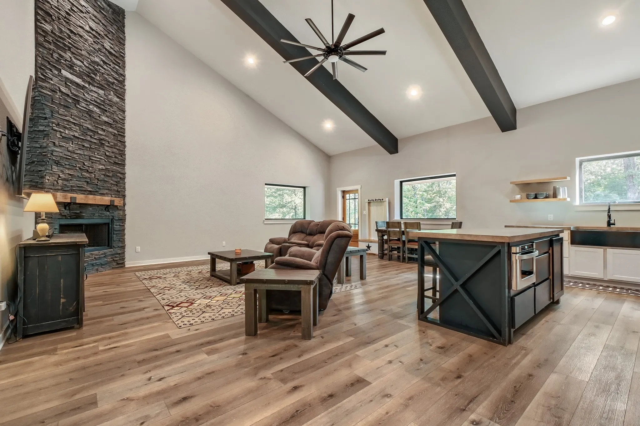 View of the open concept living, dining and kitchen, featuring vaulted ceilings with exposed beams, luxury vinyl wood flooring, floor to ceiling stone fireplace with a timber mantel and eat-in kitchen island.