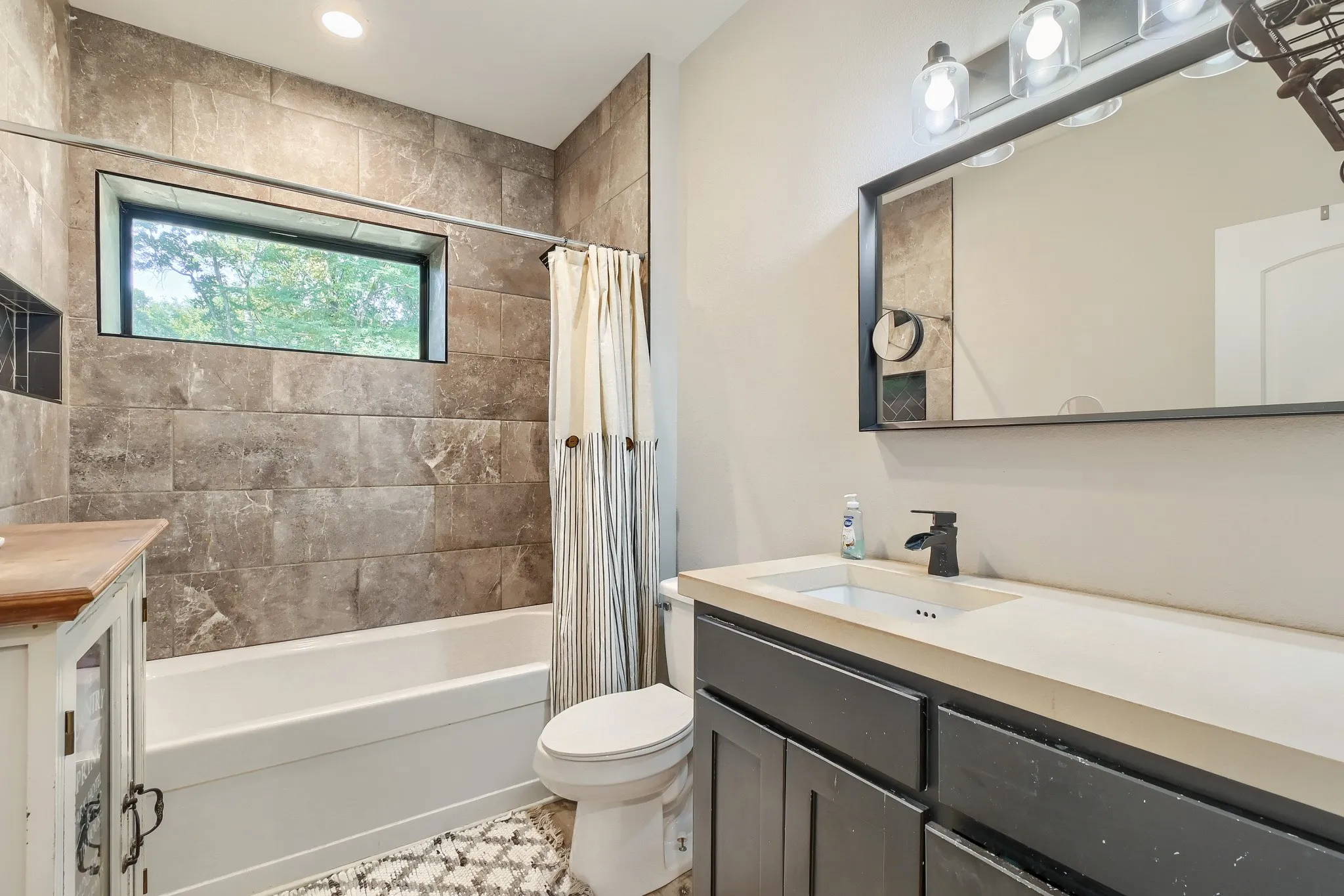 Secondary farmhouse bathroom featuring a soaking tub/shower combo with tiled walls, a horizontal window, built-in shower niche and vanity with extra storage space.