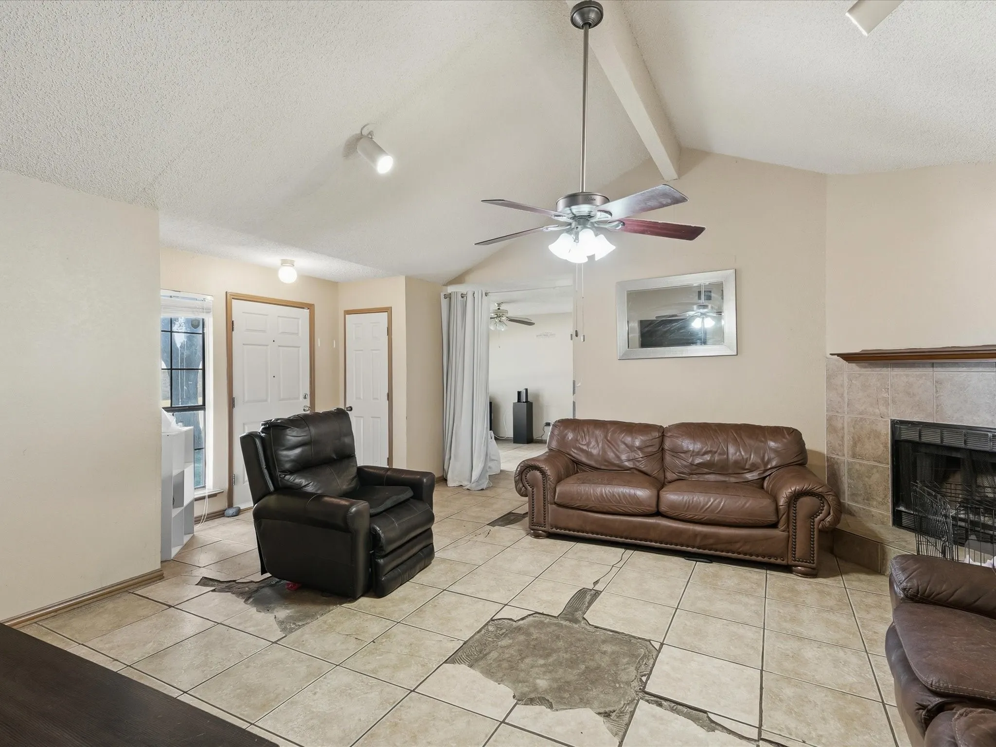 Vaulted ceiling with exposed beam, tile fireplace with mantel, ceiling fan with light fixture, tile flooring, and track lighting