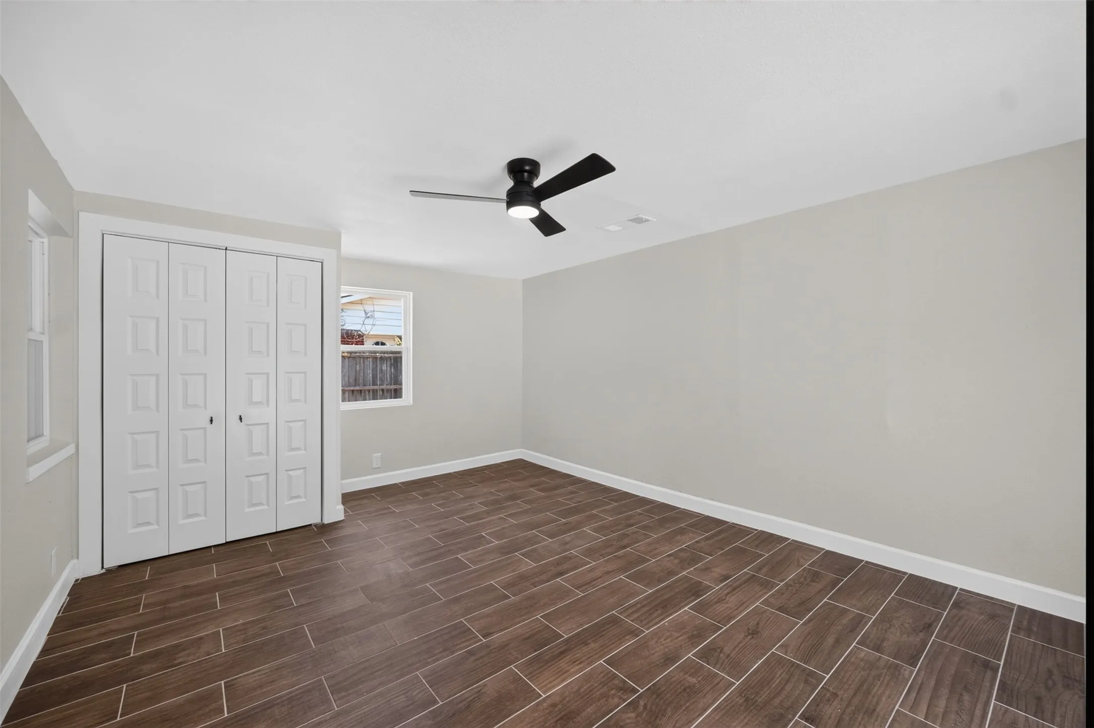Spacious room featuring wood-finish tile flooring, a modern ceiling fan with integrated lighting, and two windows providing natural illumination