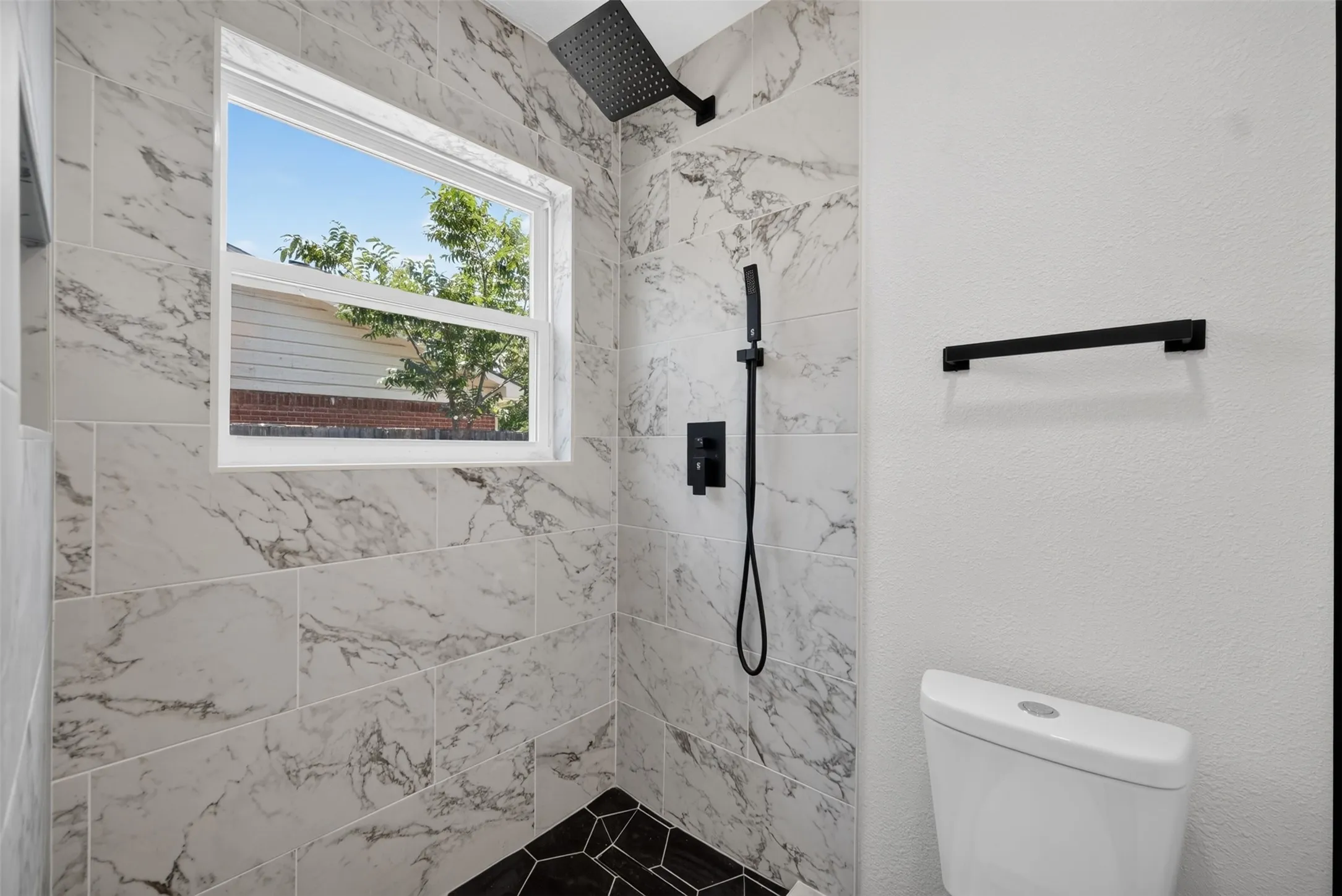Shower enclosure featuring marble-look tile walls, black hexagonal tile flooring, a matte black rain shower head, and a matte black handheld shower fixture