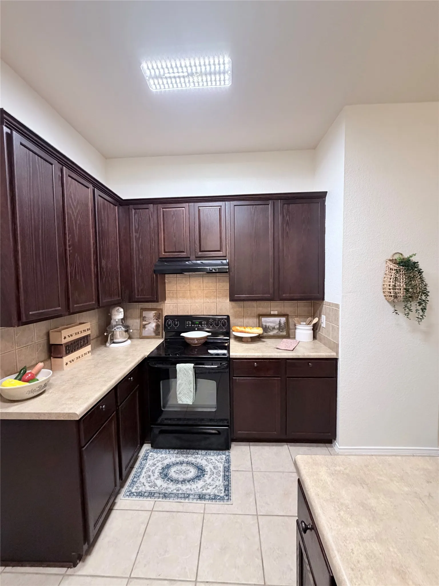 Dark wood-finish cabinetry, light-colored countertops, tile backsplash, built-in black range with overhead ventilation, and light-colored tile flooring