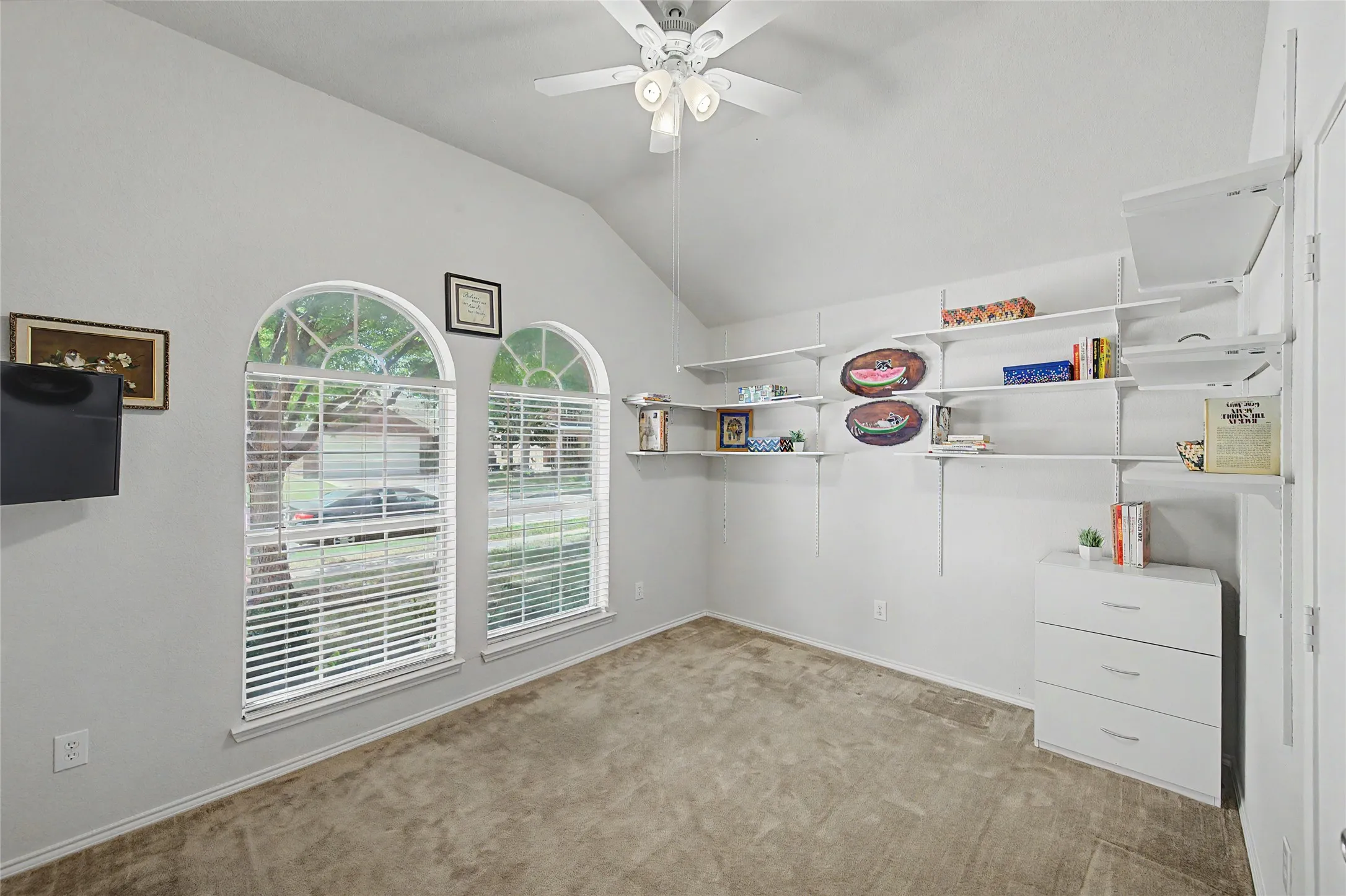 Vaulted ceiling room featuring two arched windows with blinds, track-mounted shelving, and a ceiling fan with light kit