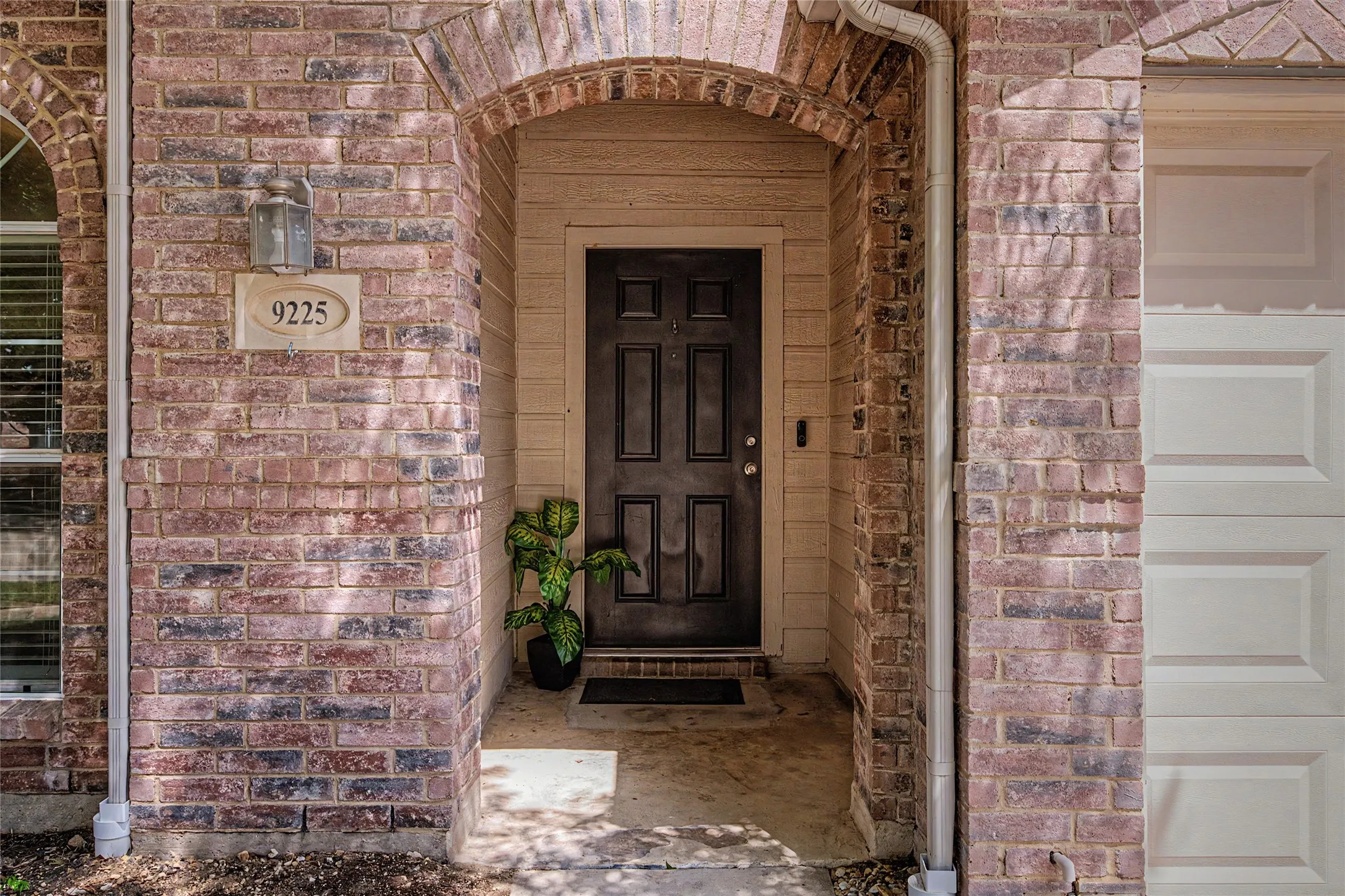 Brick facade entry with an arched portico, featuring a paneled front door, an exterior wall lantern, and a visible address plaque