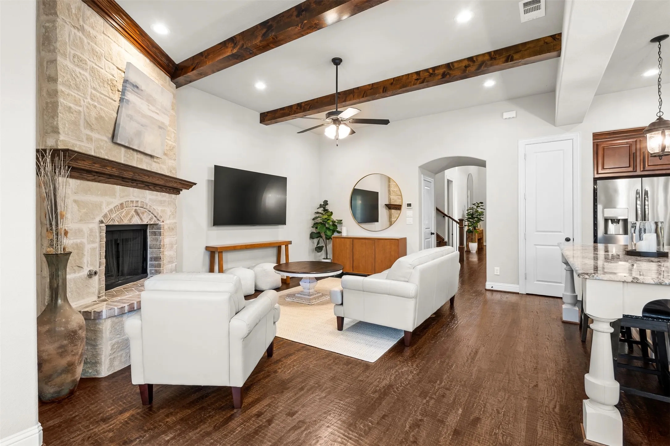 Open-concept living area featuring a stone fireplace with a brick firebox and wood mantle, wood-finish flooring, and vaulted ceilings with exposed wood beams