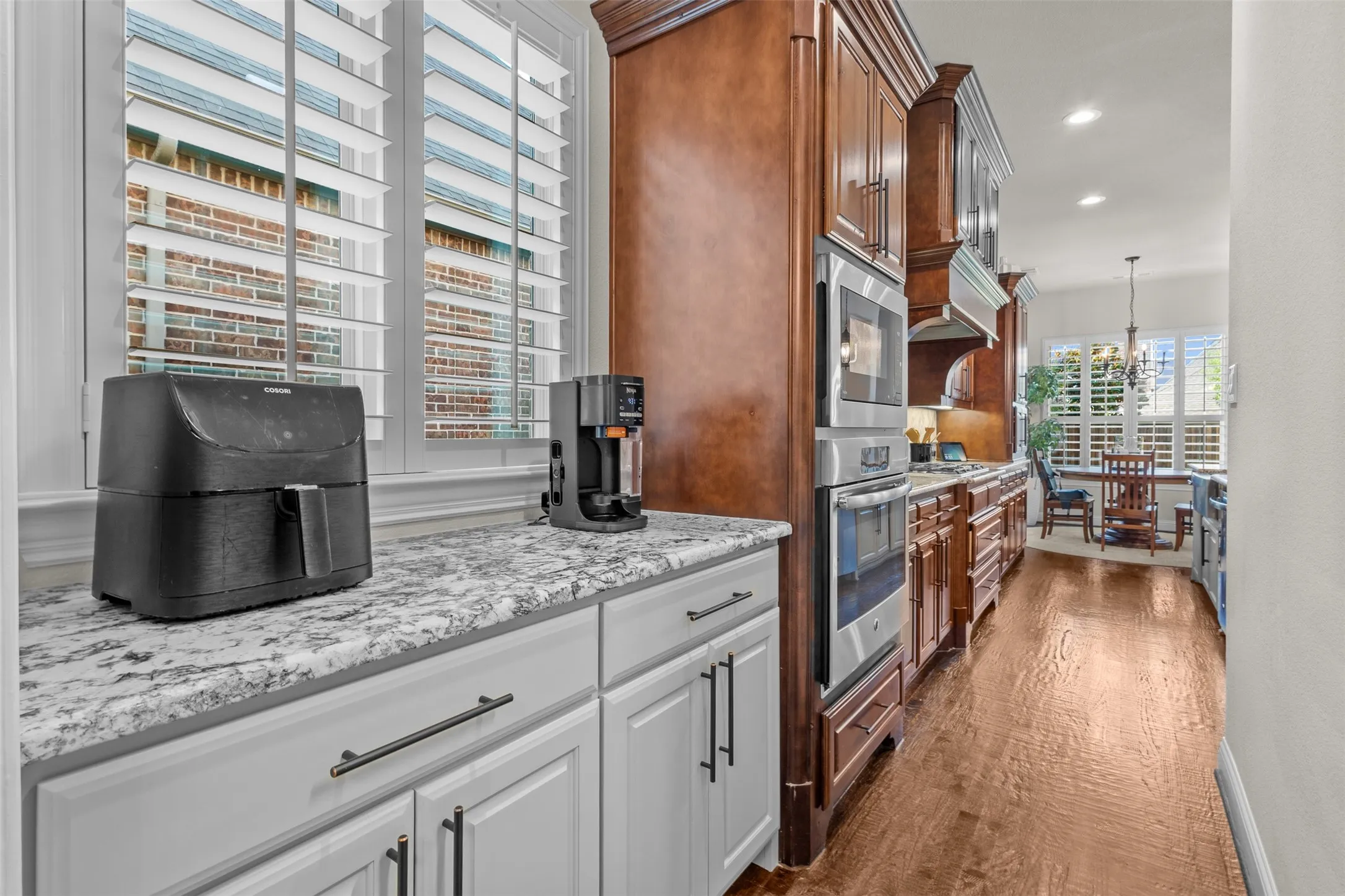 Kitchen featuring light-toned cabinetry, granite countertops, wood-finish flooring, plantation shutters, and stainless steel built-in appliances including a wall oven and microwave