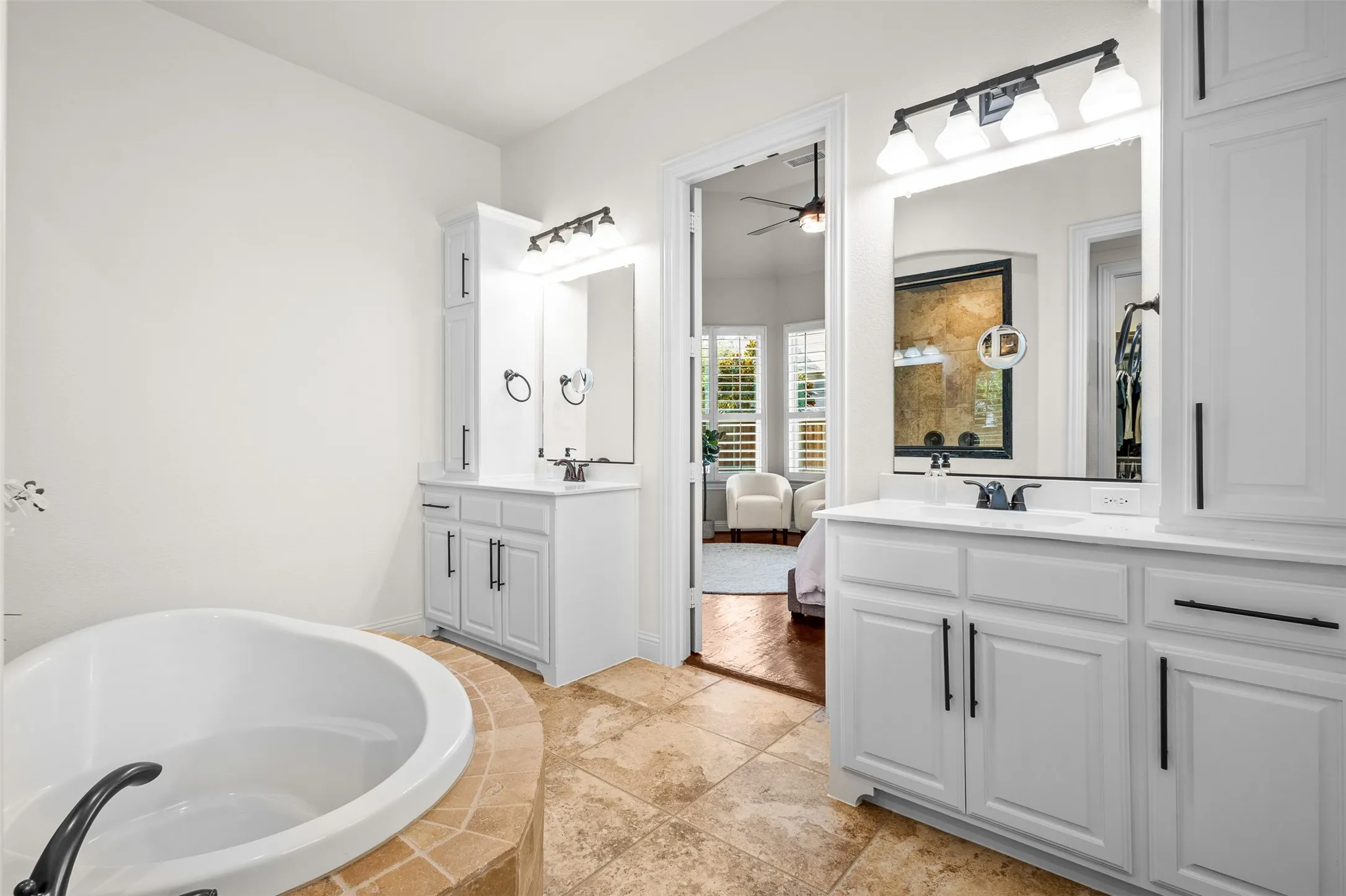 Bathroom featuring a built-in soaking tub with tile surround, dual white vanities with integrated sinks, and individual mirrors with task lighting