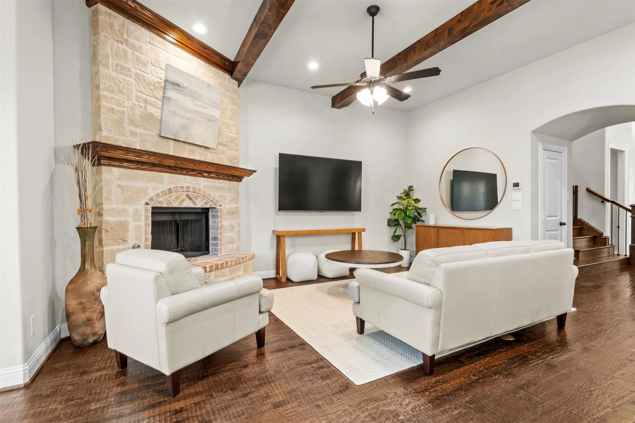 Spacious living area featuring a floor-to-ceiling stone fireplace, exposed wood ceiling beams, a ceiling fan with integrated lighting, wood-finish flooring, and a staircase with wood treads and banister