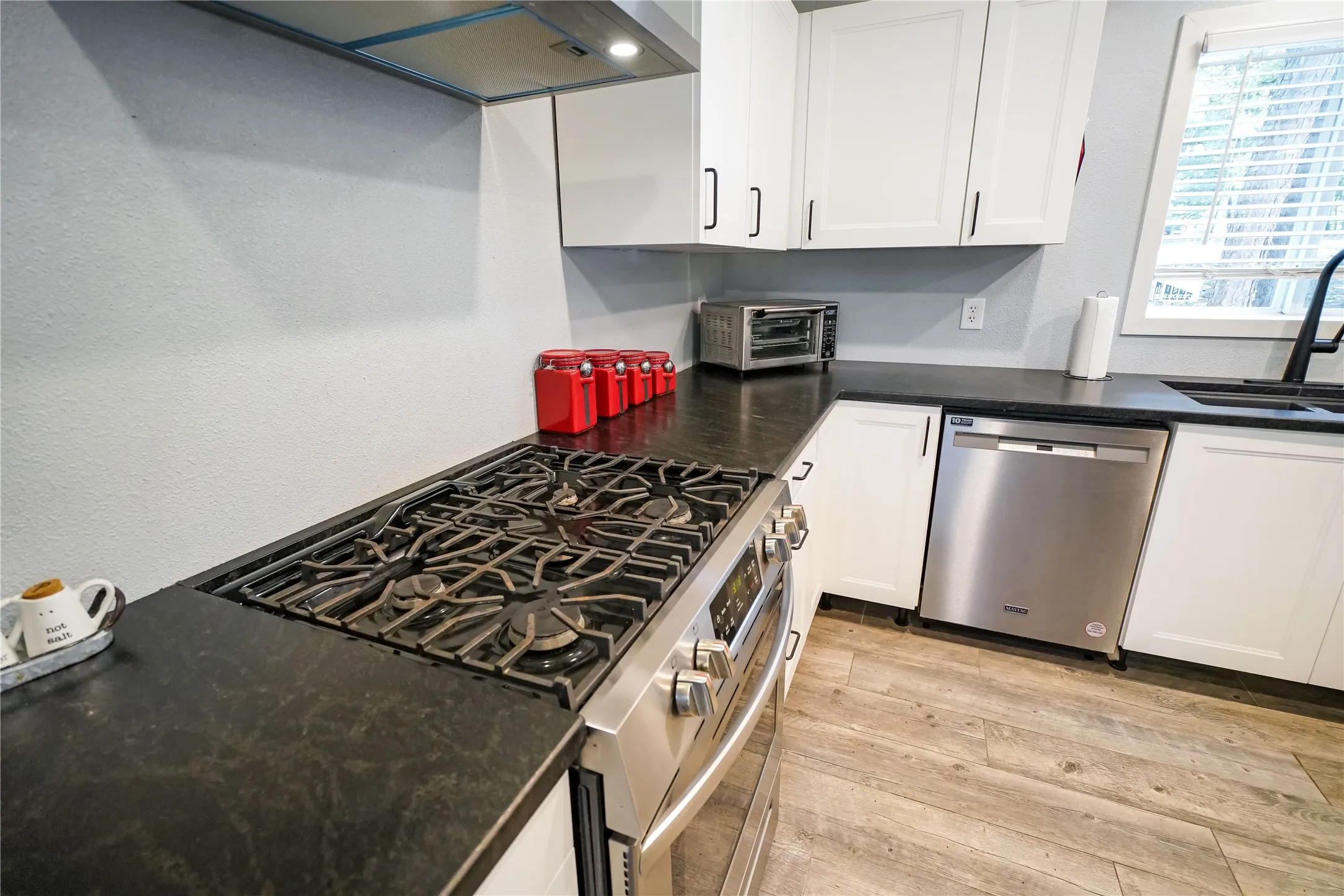 Kitchen featuring a stainless steel range with a built-in cooktop, dark countertops, white cabinetry, a stainless steel dishwasher, and wood-finish flooring