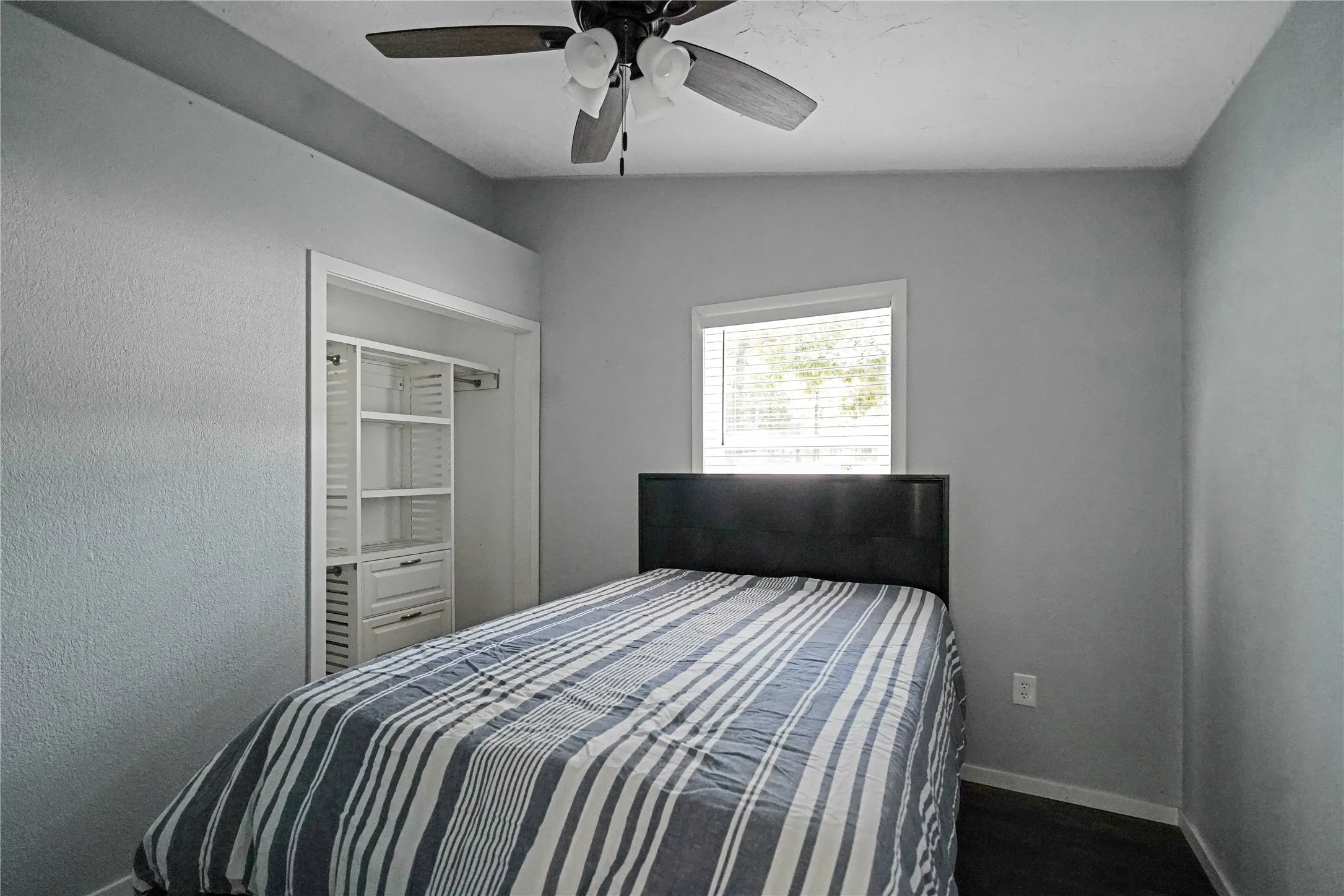 Bedroom featuring light gray walls, a window with horizontal blinds, wood-finish flooring, a ceiling fan with integrated lighting, and a built-in closet organization system