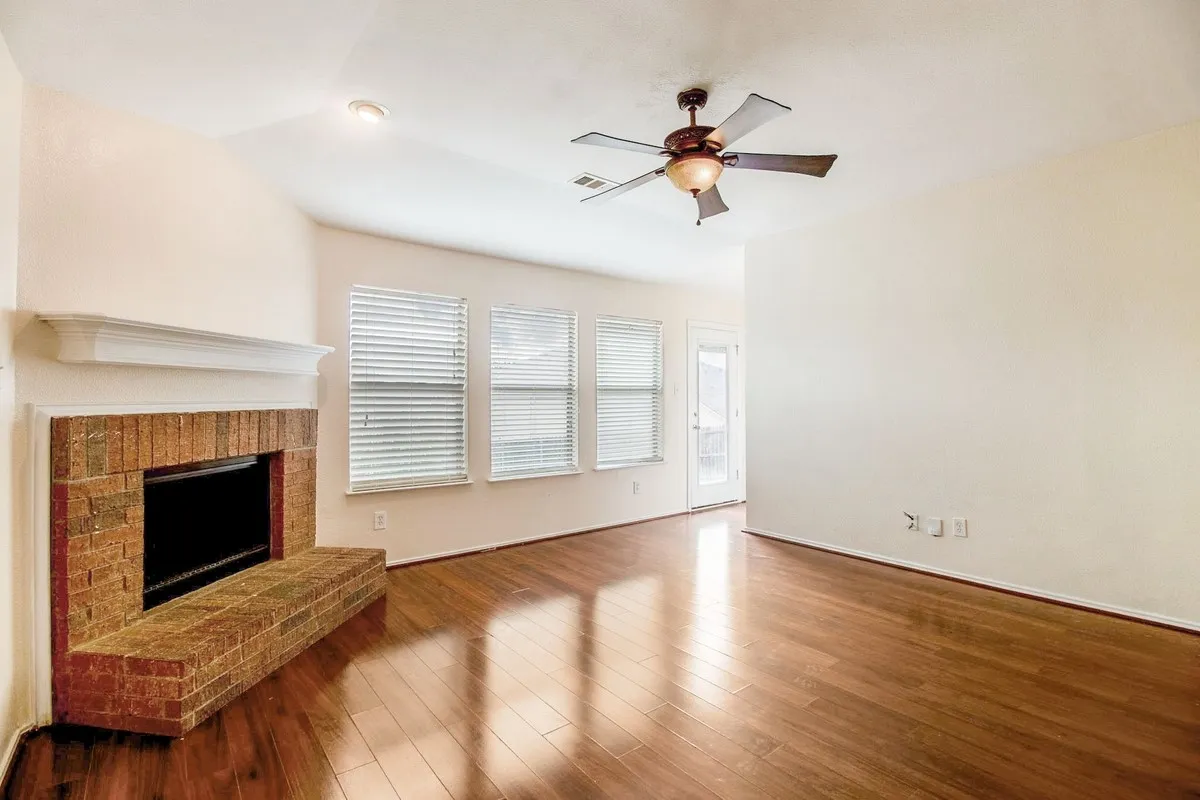 Living area featuring rich wood-finish flooring, a brick fireplace with a white mantel, and multiple windows with blinds