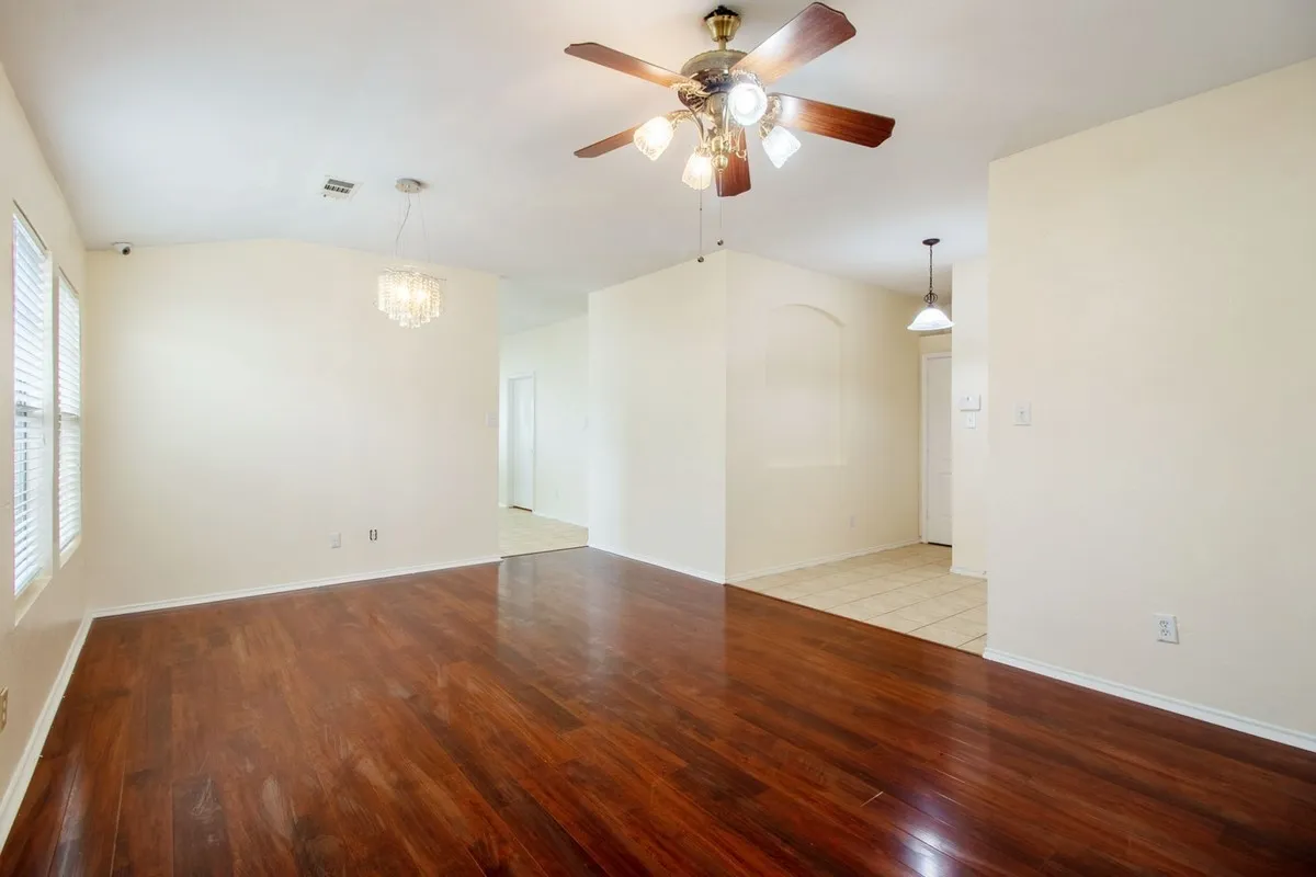 Spacious room featuring wood-finish flooring, two large windows with blinds, a ceiling fan with light kit, and a decorative chandelier