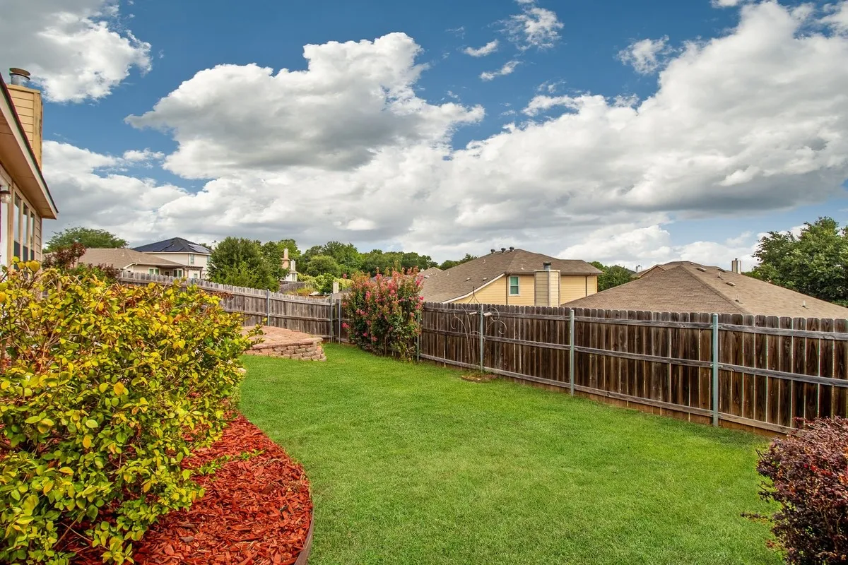 Expansive backyard featuring a lush lawn, mature perimeter landscaping, wood privacy fencing, and a built-in stone patio