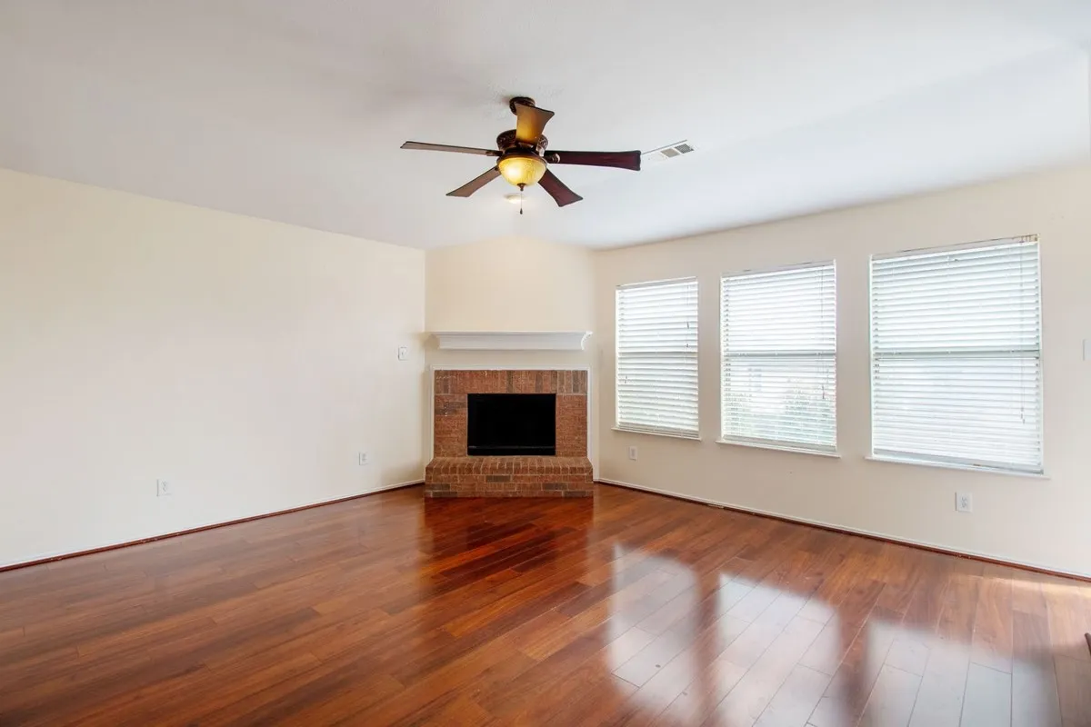 Spacious interior with wood-finish flooring, a brick fireplace with mantel, and three windows featuring blinds