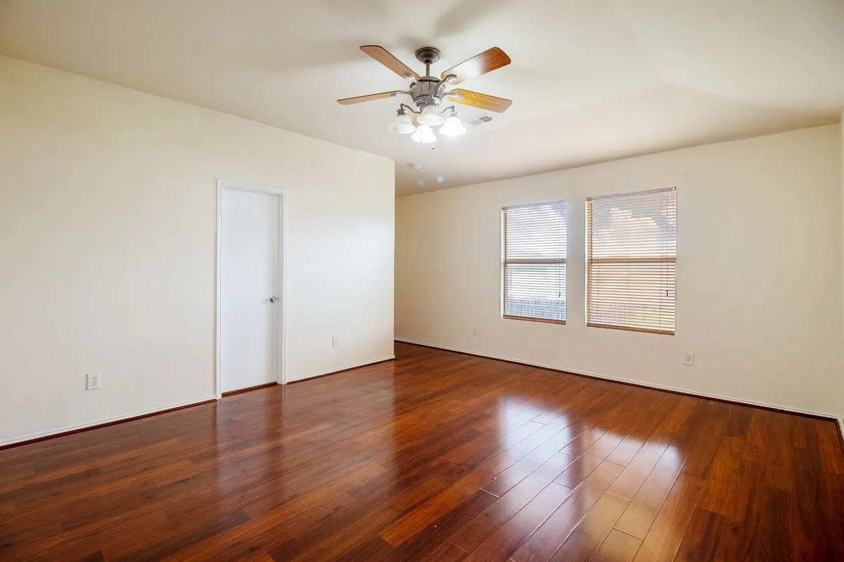 Spacious room featuring wood-finish flooring, light-toned walls, a ceiling fan with integrated lighting, and two windows with horizontal blinds