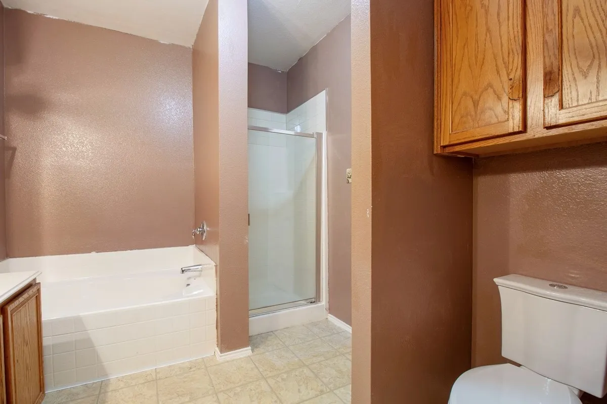 Bathroom featuring a built-in bathtub, separate shower with a glass enclosure, light-toned tile flooring, wood-finish cabinetry, and a white toilet