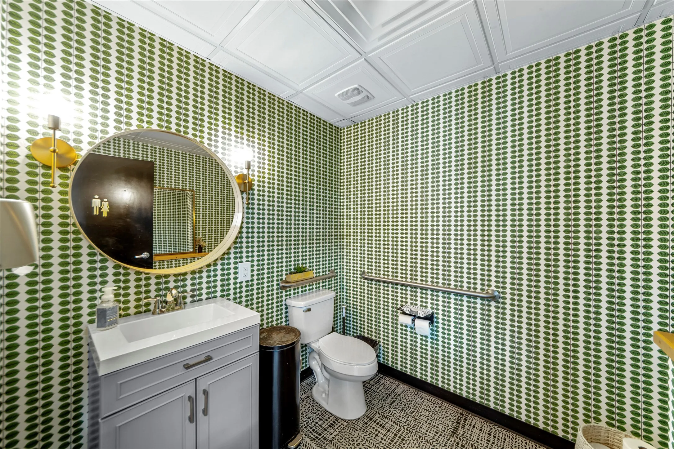 Bathroom featuring green patterned wallpaper, a white vanity with a rectangular sink, a gold-framed circular mirror, and twin wall-mounted sconces
