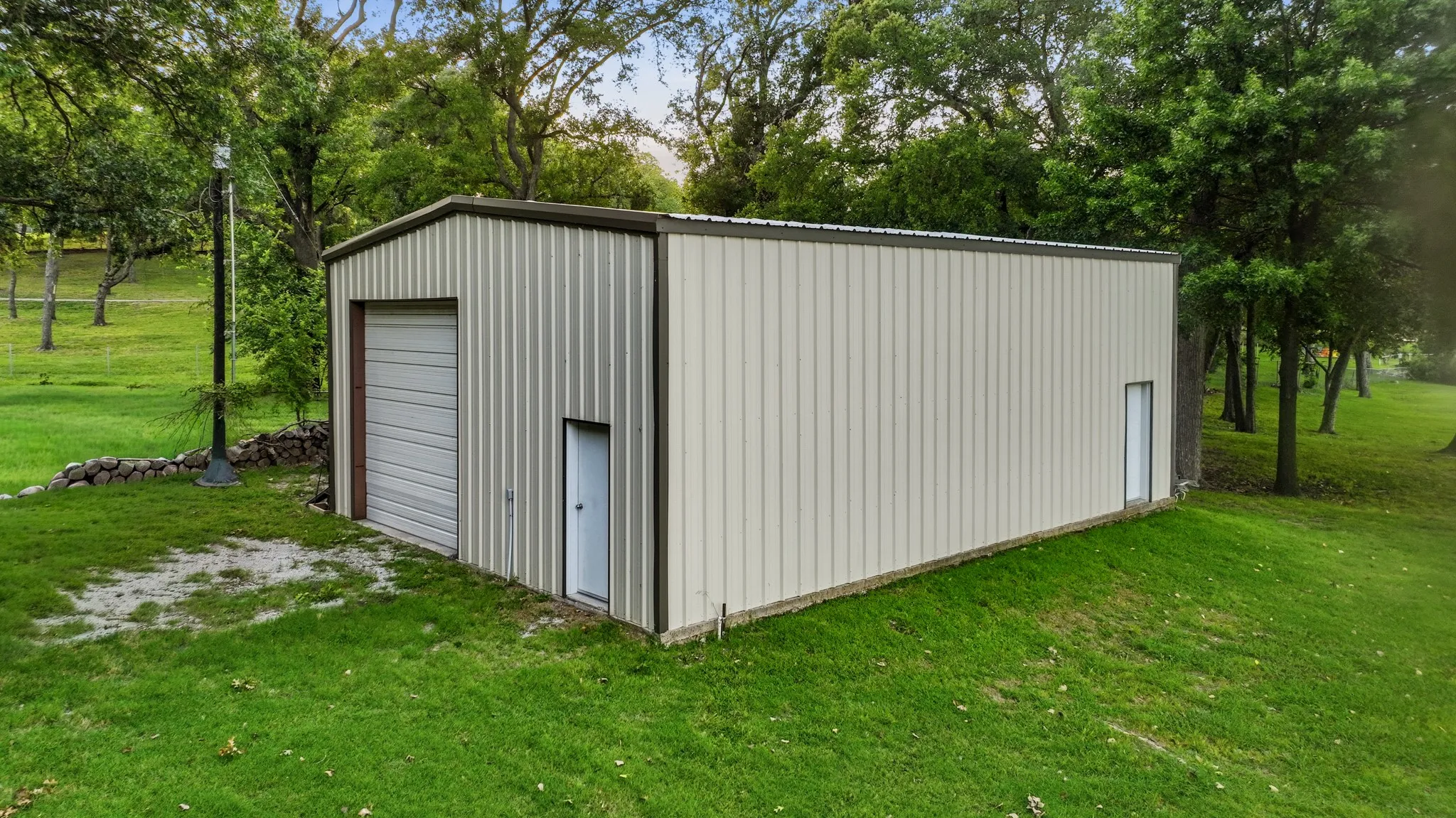 Metal outbuilding featuring a roll-up garage door and two side entry doors, set on a manicured lawn with mature trees