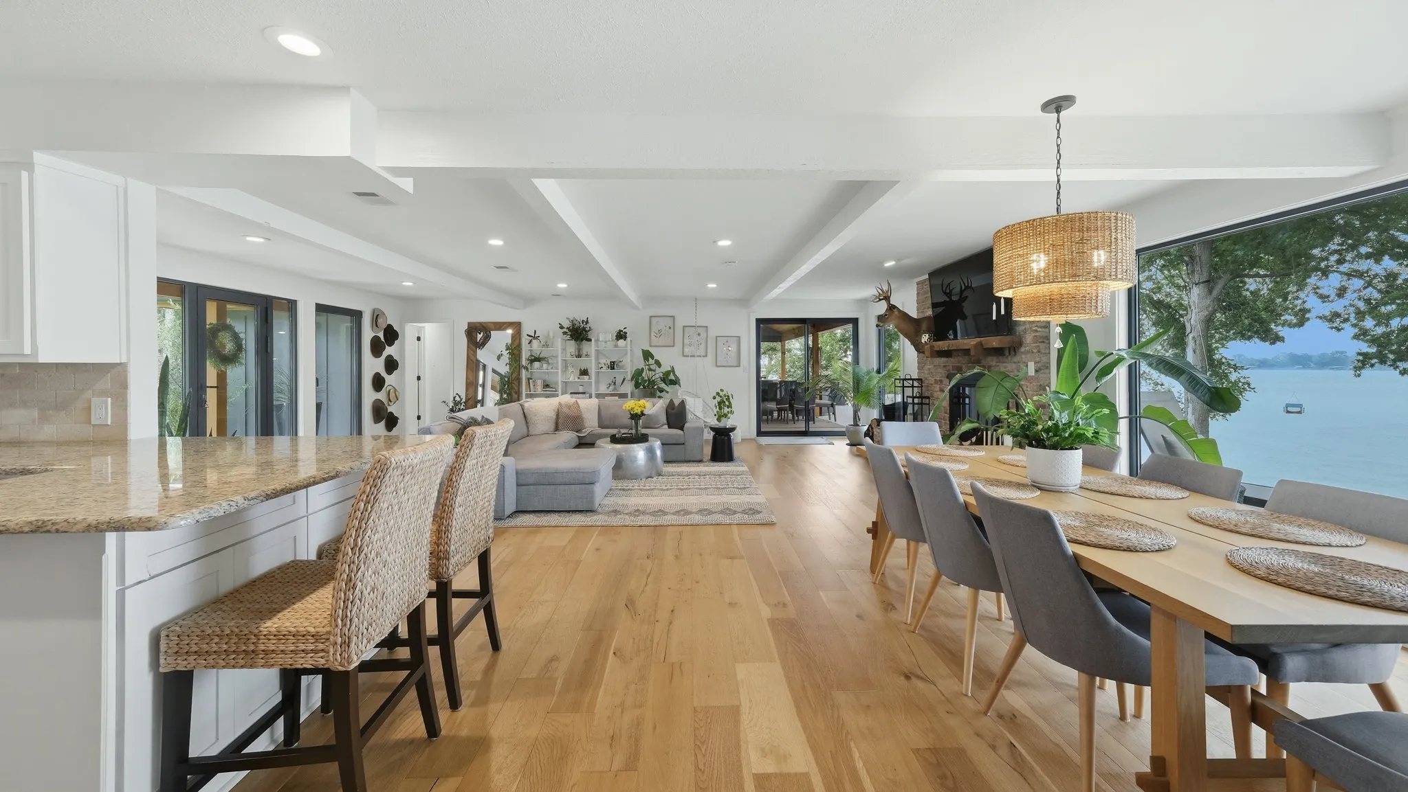 Open-concept living space featuring wood-finish flooring, a contemporary dining area with a woven chandelier, a stone fireplace, and expansive windows offering water frontage