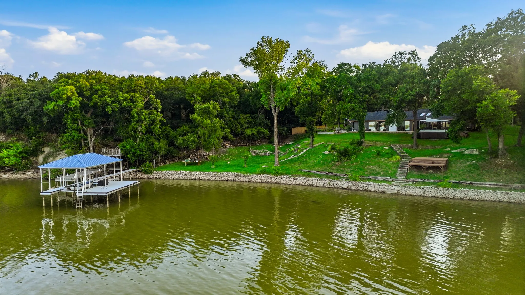 Waterfront property featuring a private boat dock with a blue roof, a two-story house with a covered patio, a terraced lawn, and mature trees
