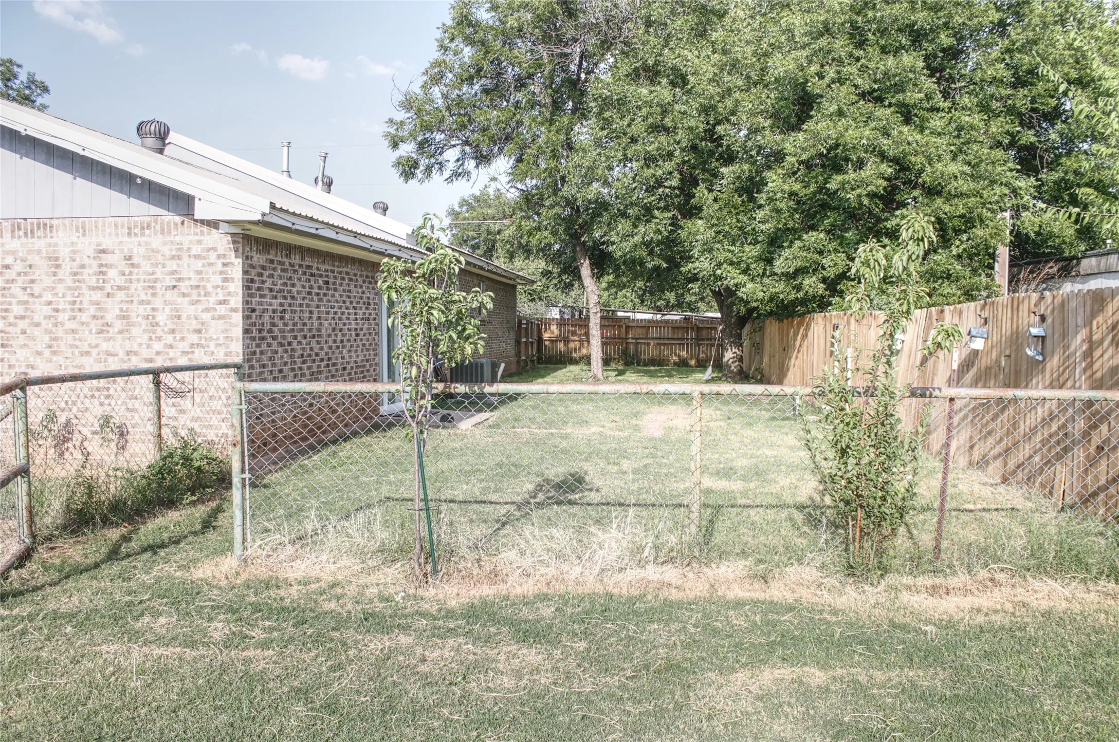 Expansive backyard featuring a partial chain-link fence, a partial wood privacy fence, mature trees, and a brick exterior structure